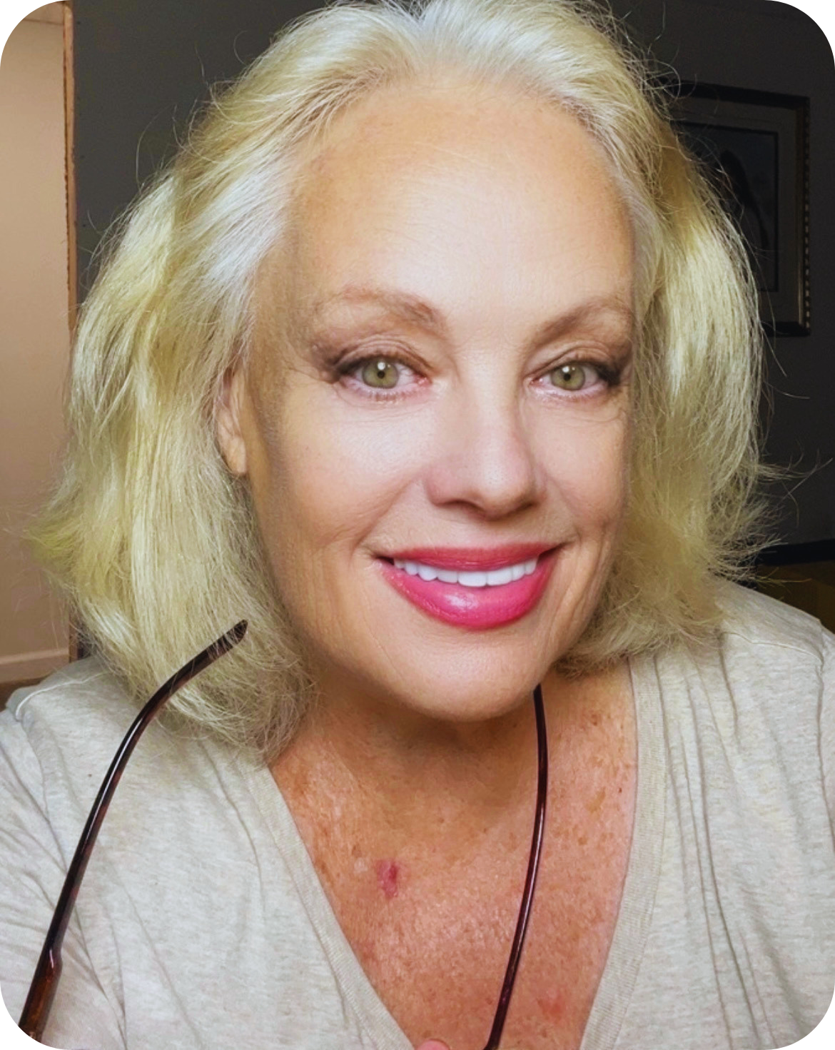 A middle-aged woman with blonde hair, green eyes, pink lipstick, smiling, holding glasses, wearing a light-colored top, in an indoor setting.