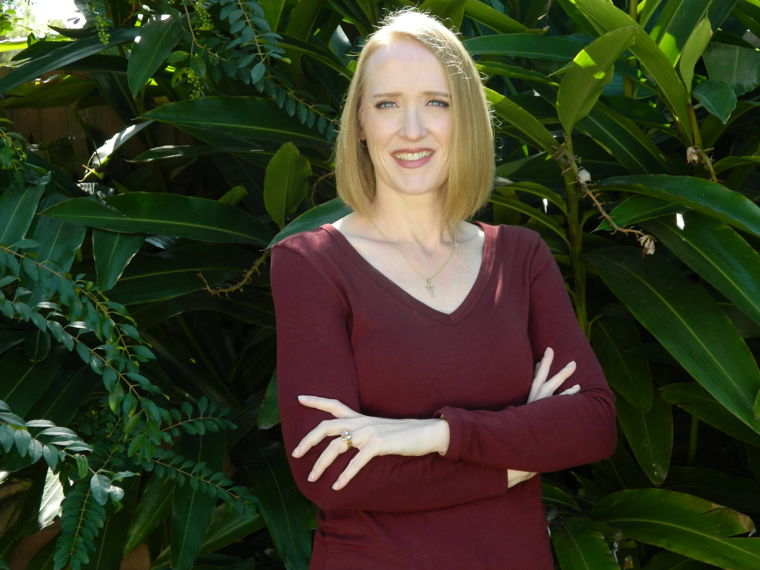 A woman with blonde hair, wearing a burgundy long-sleeve shirt, standing outdoors in front of green leafy plants.