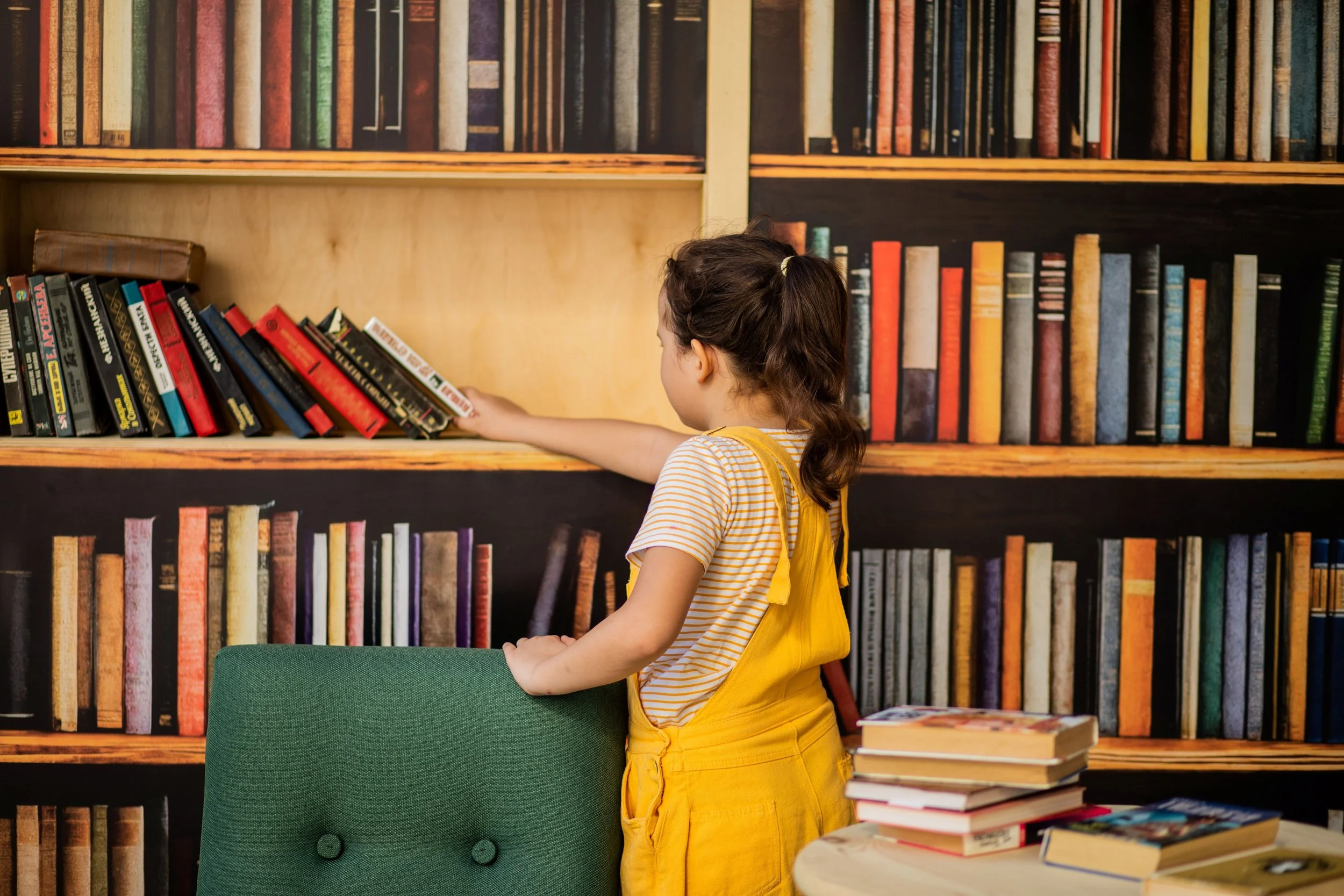 young girl in yellow overalls and a striped t-shirt, reaching for a book from a large bookshelf