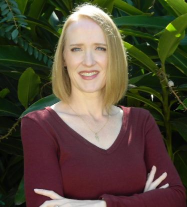 A woman with blonde hair, wearing a burgundy long-sleeve shirt, standing outdoors in front of green leafy plants.
