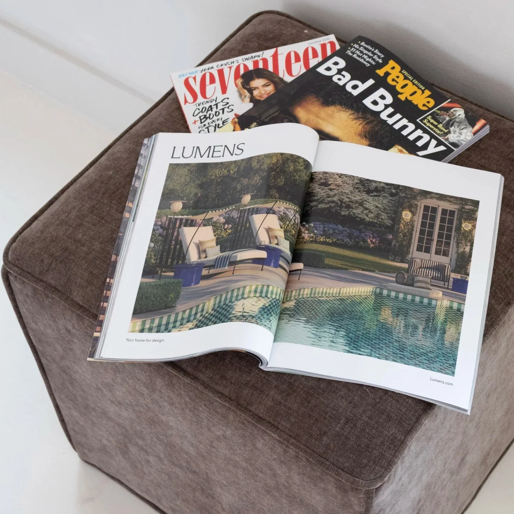 Open magazine on a brown fabric ottoman displays a two-page spread of a garden and pool scene at night, with the heading 'Lumens' and the tagline 'Your home for design'.
