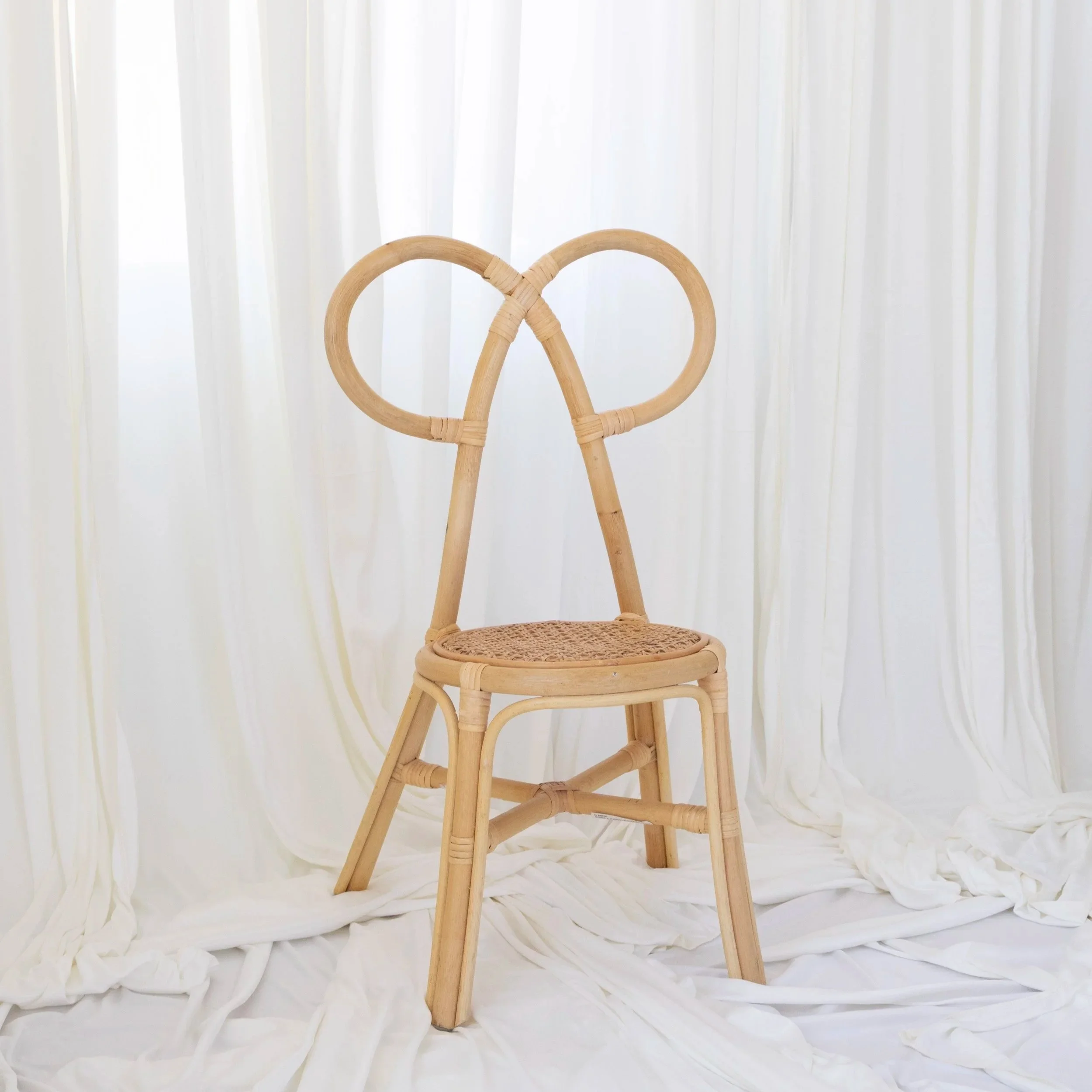 A rattan chair with a heart-shaped backrest set against white curtains and draped fabric.