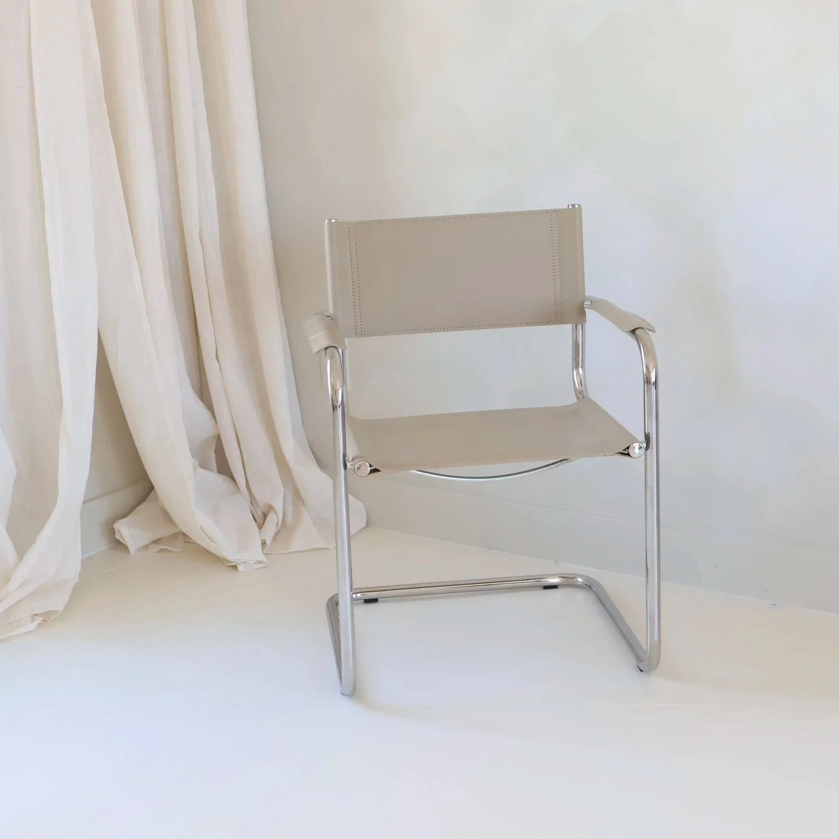 An empty beige chair with metal armrests against a plain white wall, with white curtains hanging nearby.