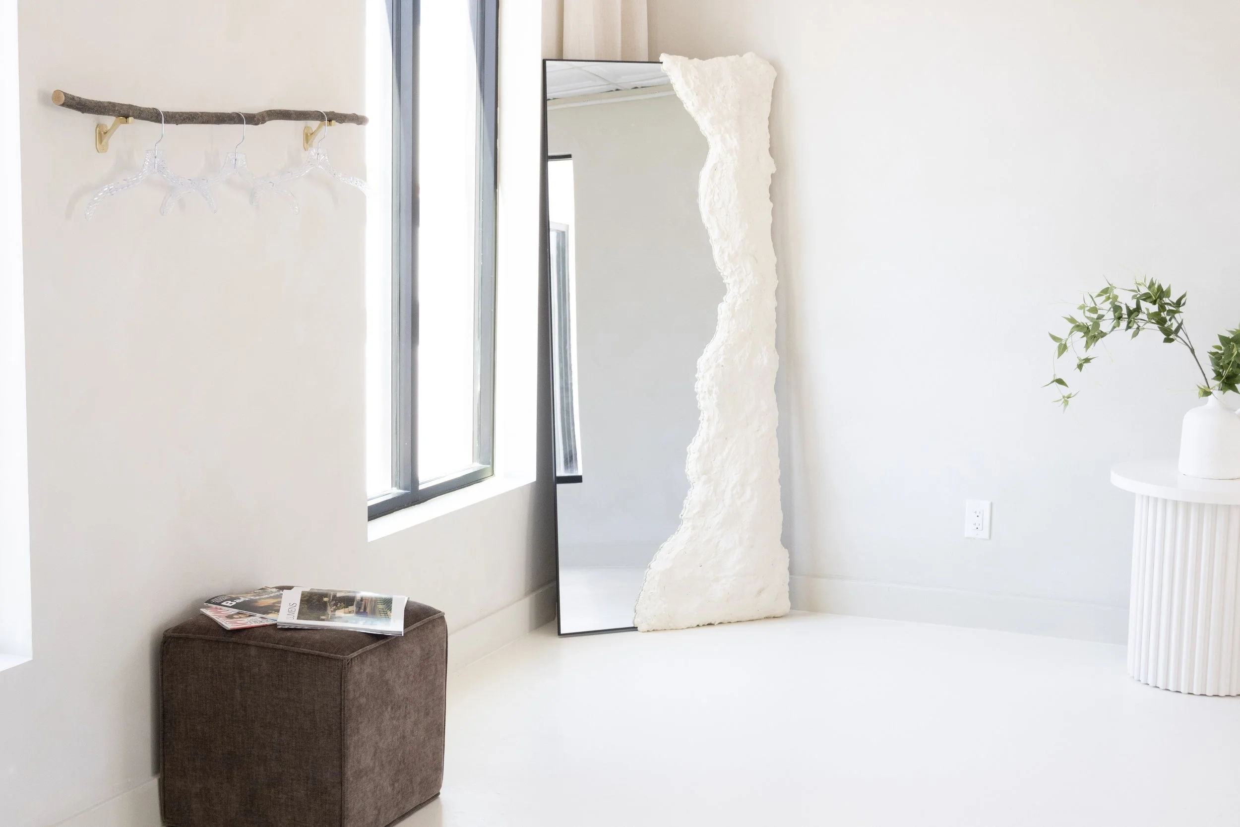 Minimalist interior with a leaning mirror, a small brown ottoman with magazines on top, a white wall decorated with a natural wood branch holding clear hangers, a window letting in bright natural light, and a green plant in a white pot.