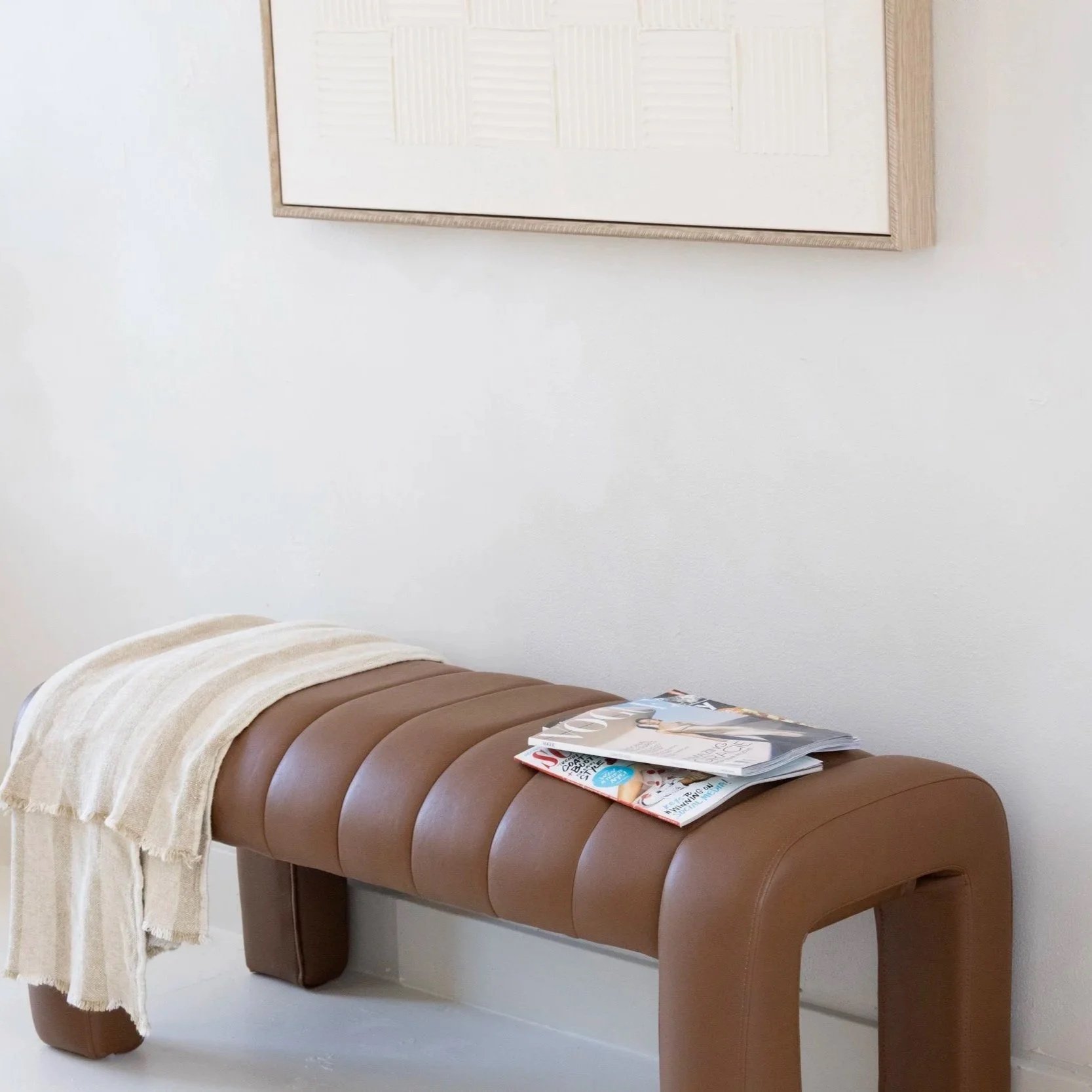 A brown massage table with a cream-colored towel and magazines on top, situated against a white wall with a framed picture.