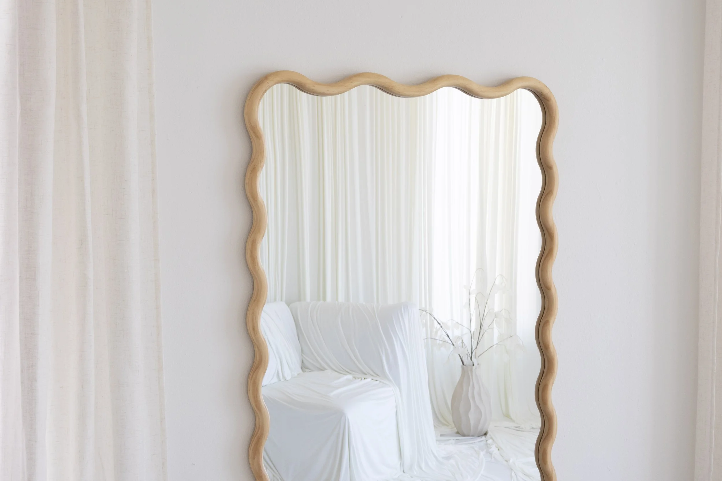 A mirror with a wavy wooden frame hung on a white wall, with part of a white bed and light curtains visible in the reflection.