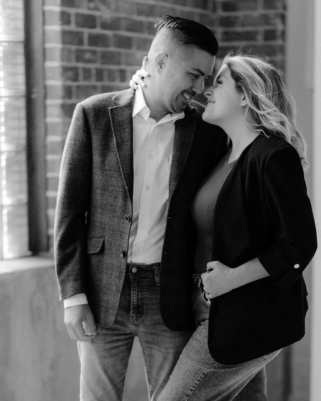 A black and white photo of a man and woman laughing and leaning forehead to forehead, with the man holding the woman's waist, against a brick wall background.