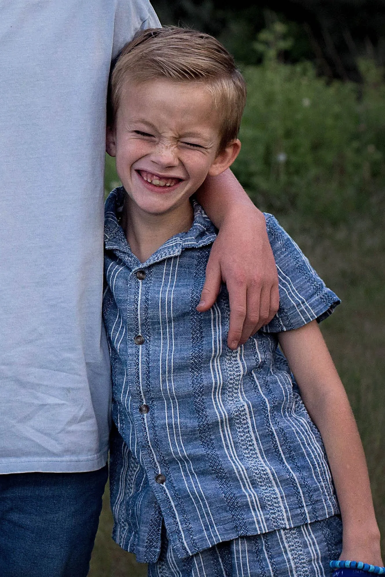 A young boy with a big smile, squinting his eyes, is seen standing outdoors next to a person who is partially visible. The boy is wearing a blue striped button-up shirt. The background is a blurred green outdoor setting.
