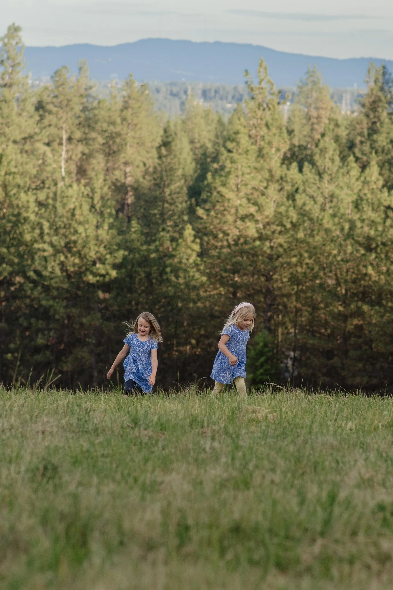 Two young girls in blue dresses playing and walking in a grassy field with trees and mountains in the background in eastern washington