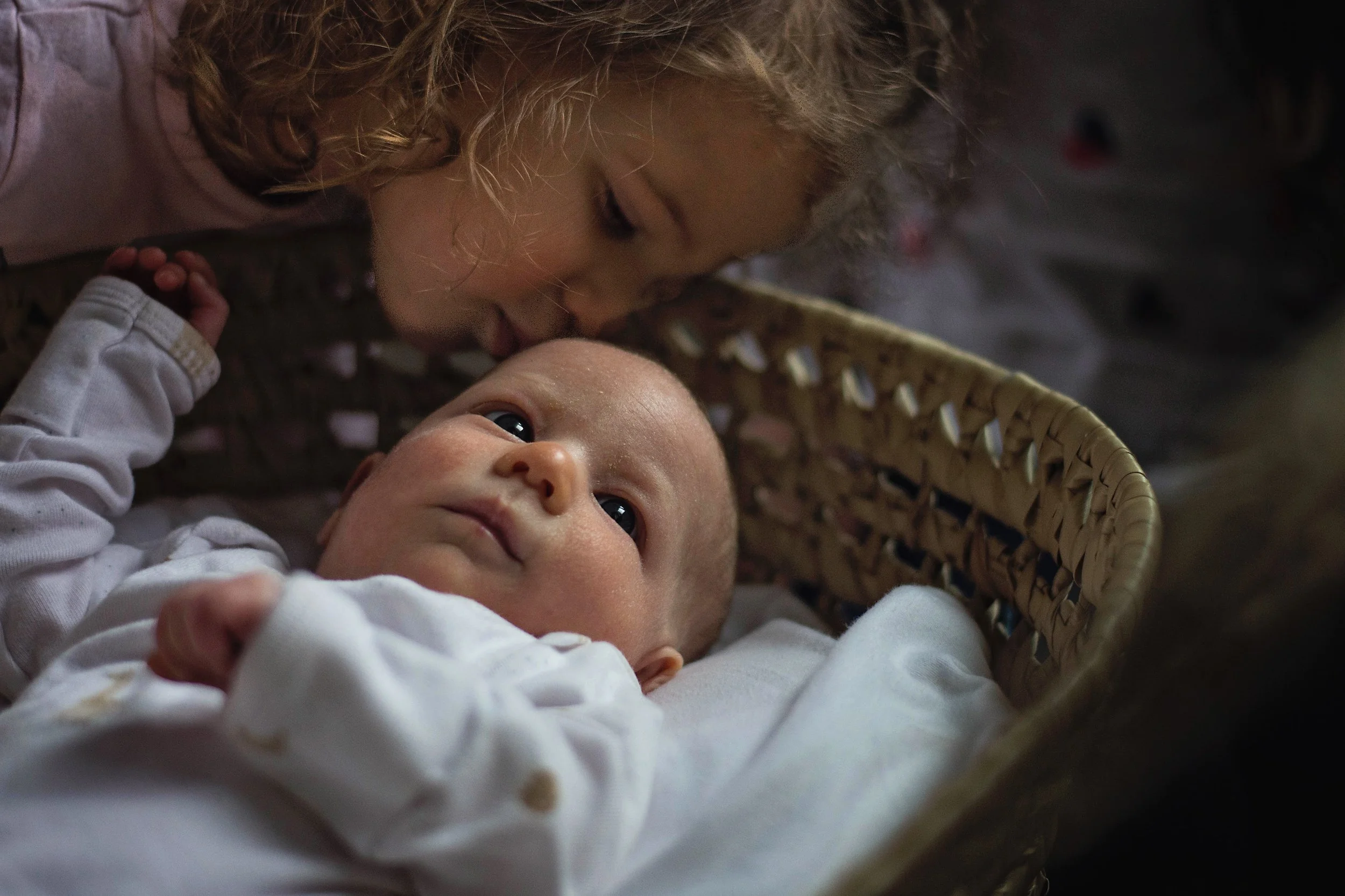 a professional photo of A young girl with curly hair leaning over a baby lying in a woven basket, gazing at the baby who looks up at her.