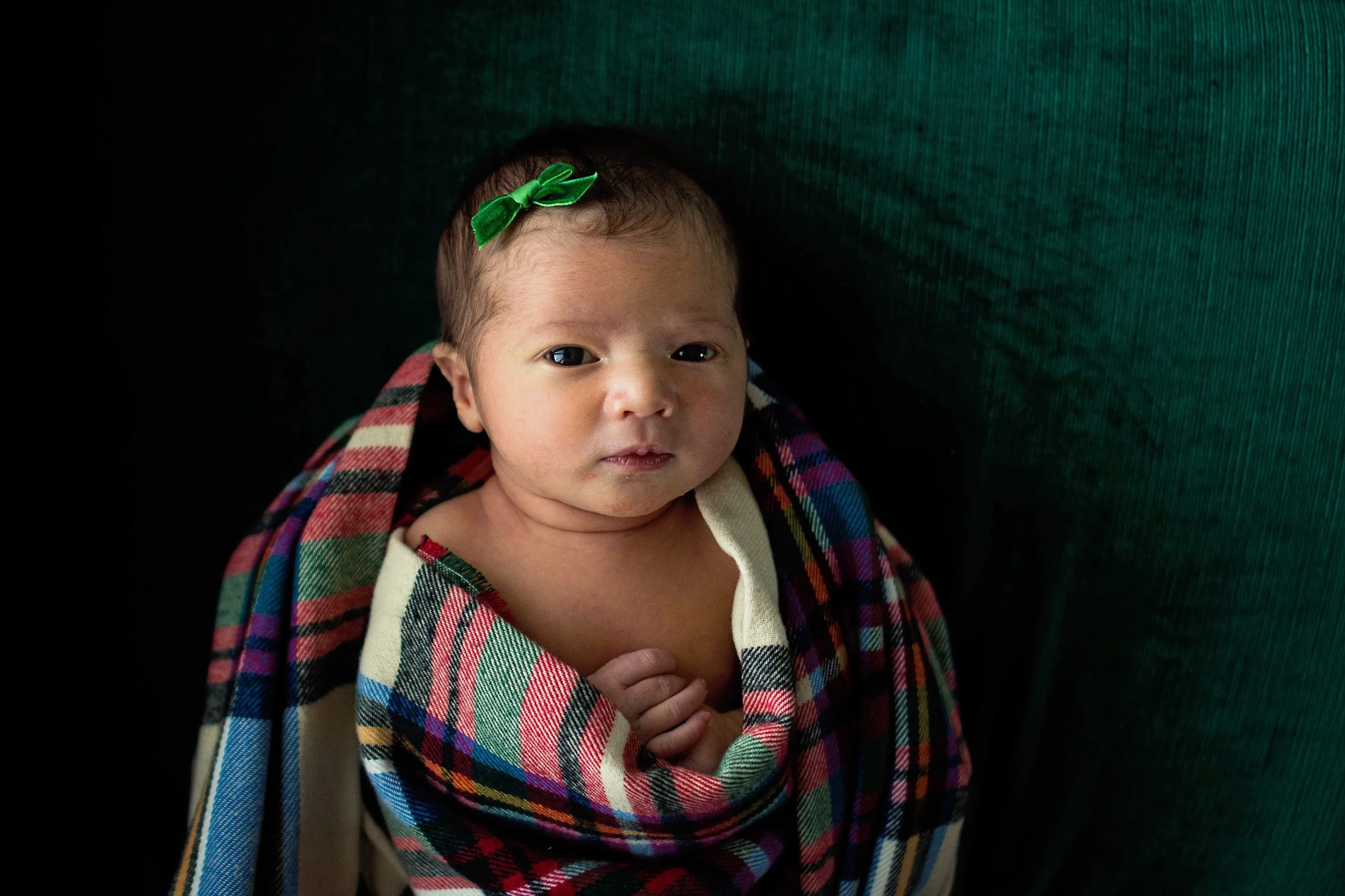 Close-up of a baby with a green bow in hair, wrapped in a colorful plaid blanket, against a dark background.