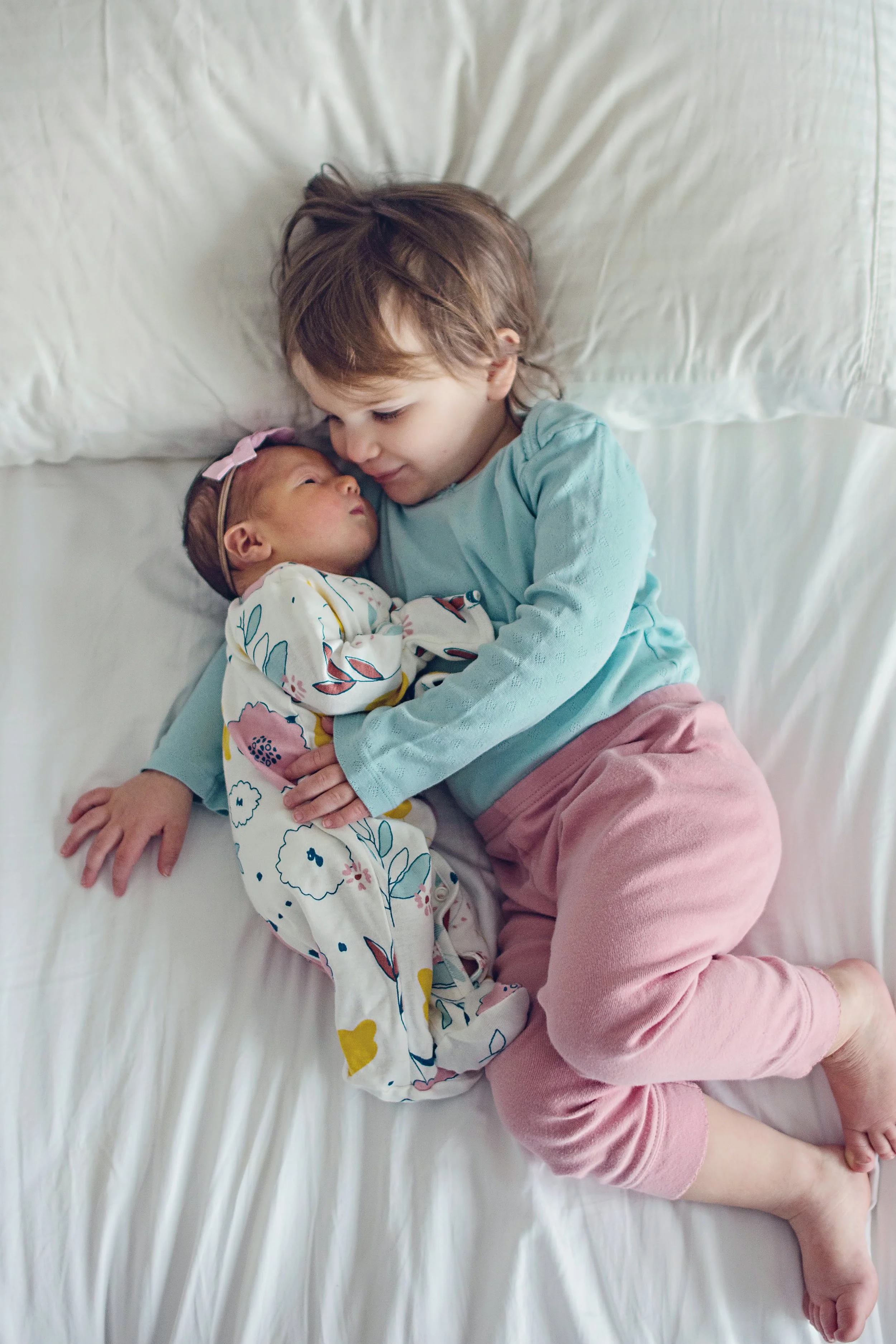 A young child lying on a bed holding a newborn baby girl, both looking at each other affectionately.