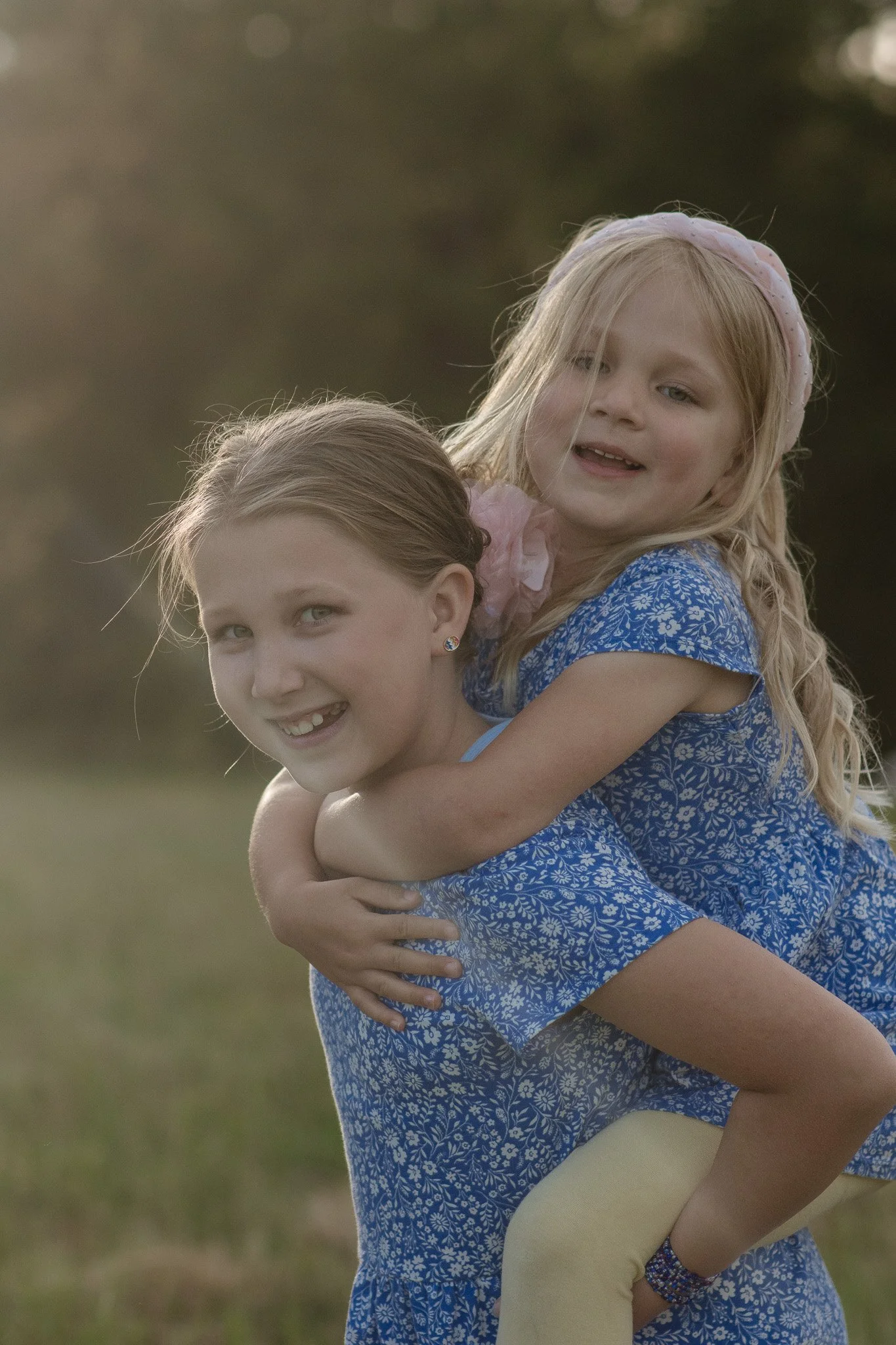 Two young girls, one with blonde hair and the other with brown hair, enjoying an outdoor moment together, with the blonde girl on the brown-haired girl's back, both smiling, in a natural setting.