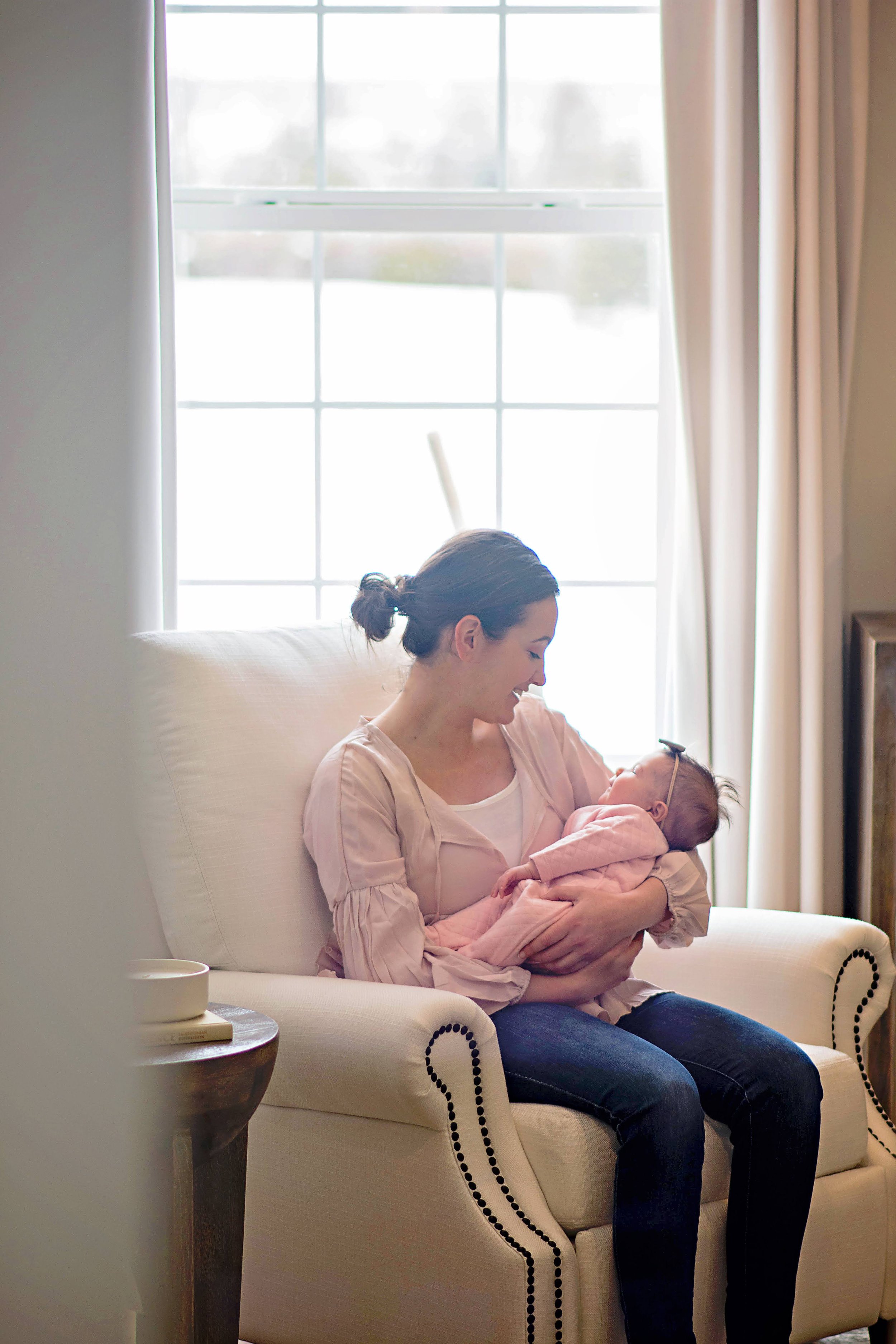 A woman sitting on a cream-colored couch holding a baby, both smiling, near a large window with curtains, in a bright room.