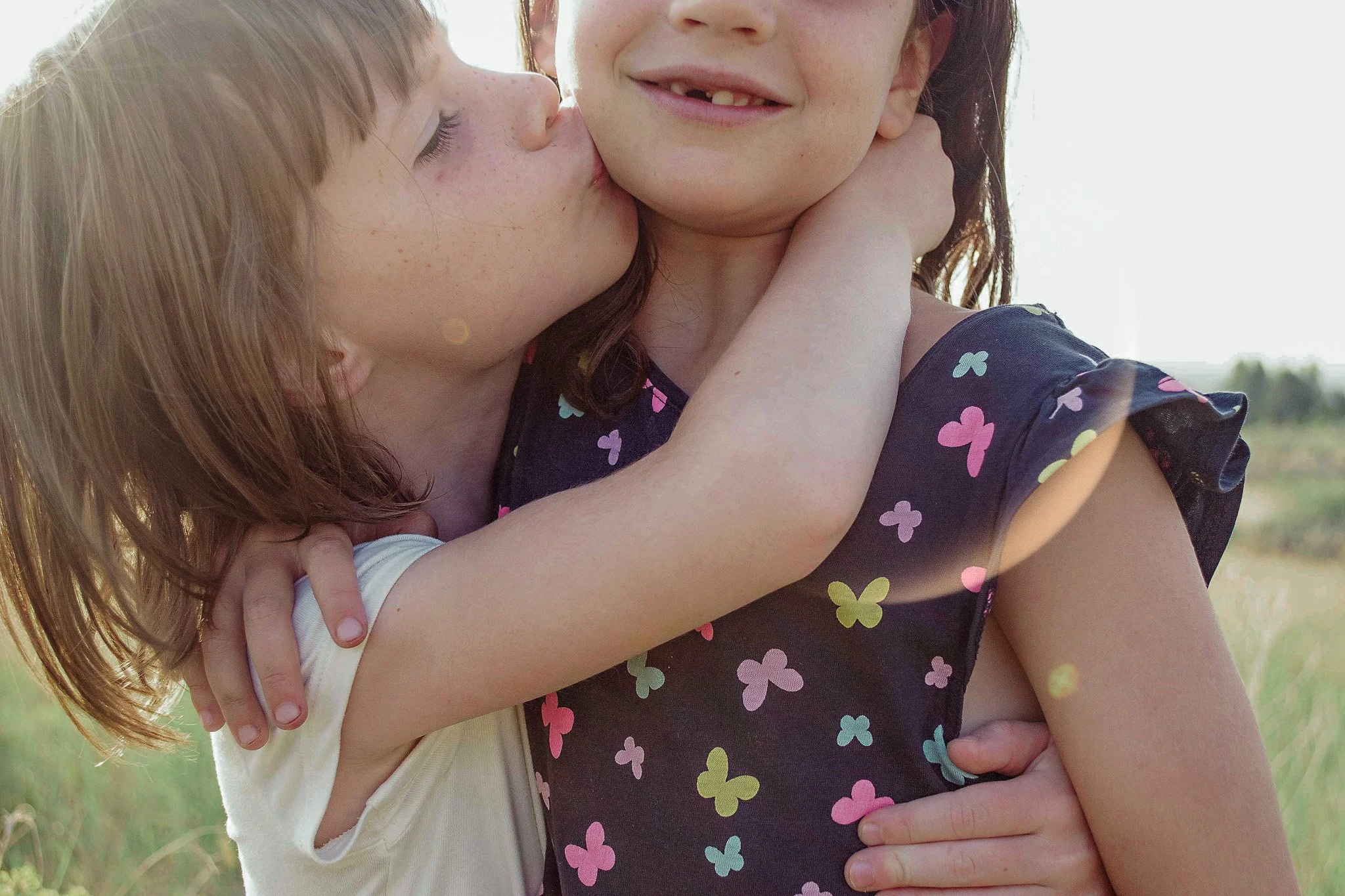 Two young girls embracing outdoors, one kissing the other's cheek, with a blurred green landscape in the background.
