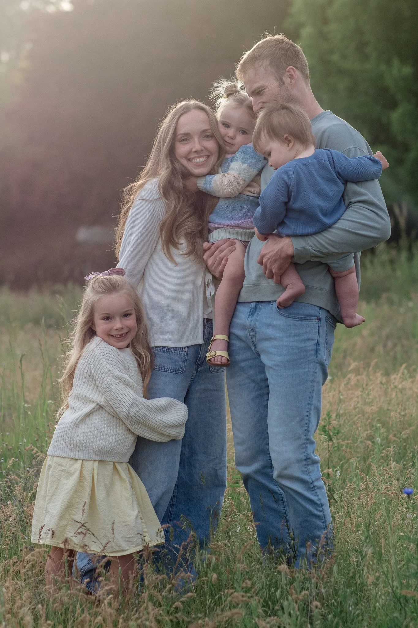 A family of five, including a mother, father, and three young children, poses together outdoors in a grassy field during the late afternoon or early evening. The mother has long, wavy hair and is smiling, while the father is holding two children, one