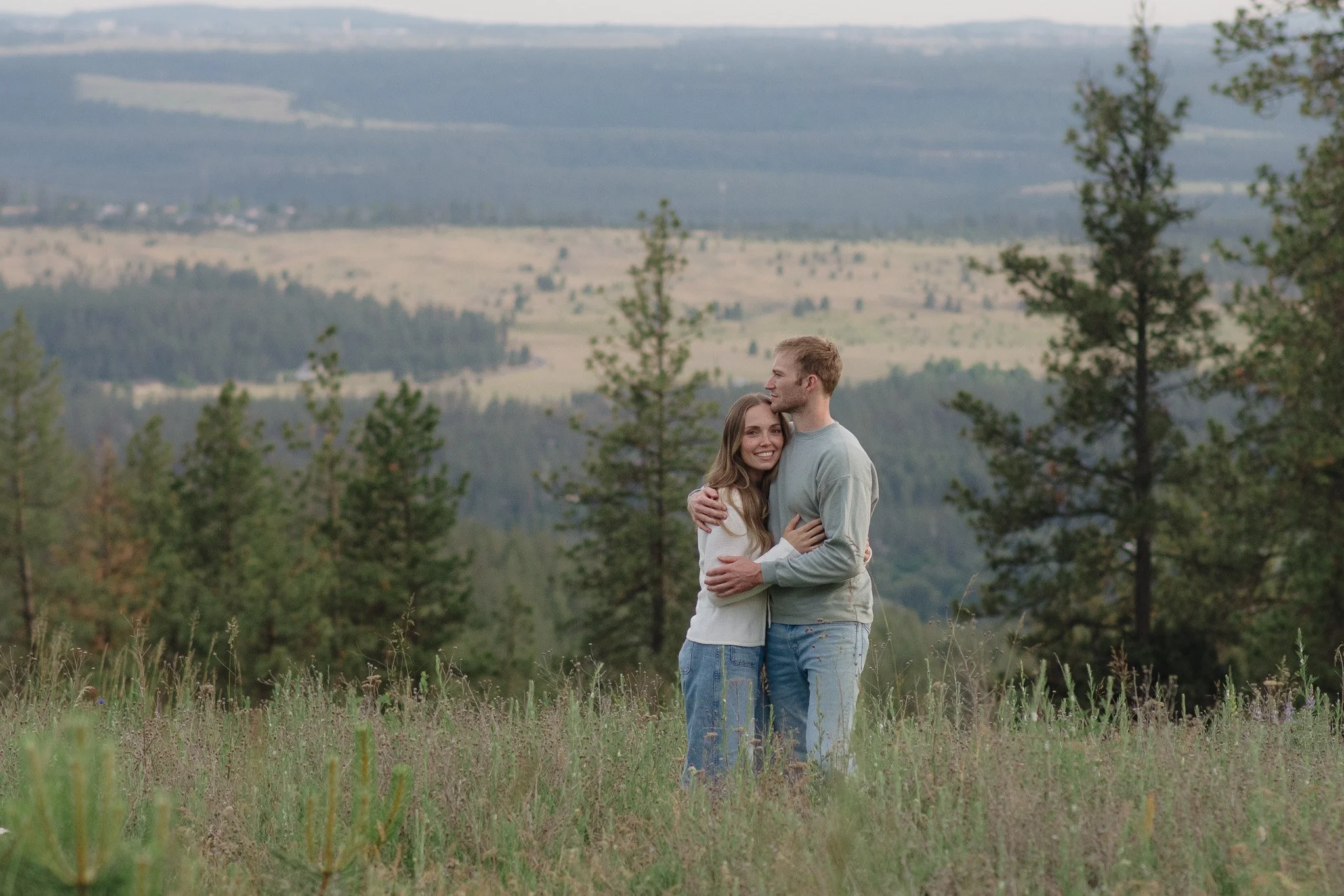A young couple hugging in a grassy field with trees, hills, and a cloudy sky in the background.
