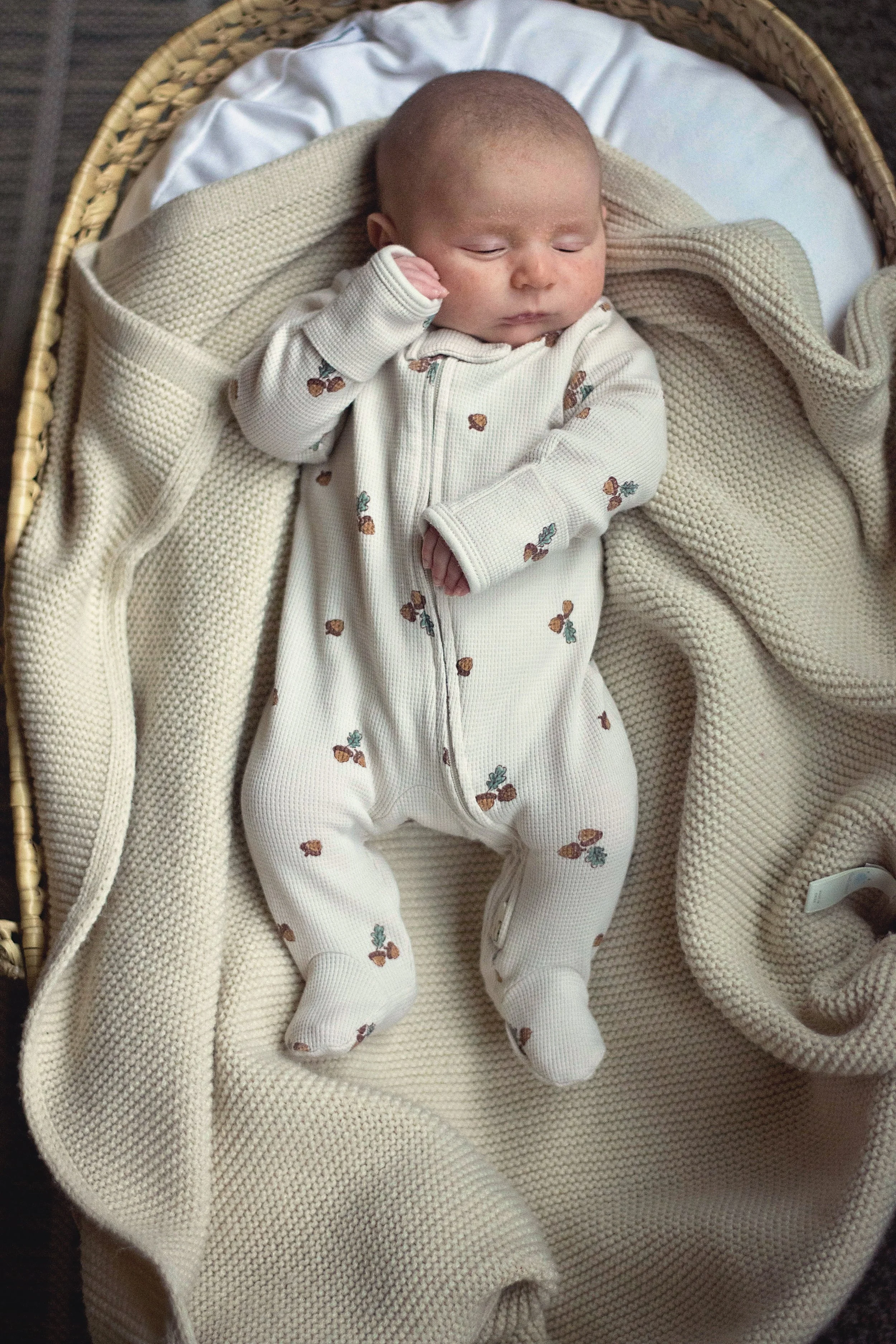 A lifestyle photograph of a baby lying in a basket on a cozy blanket, wearing a cream-colored onesie with a small floral pattern, and snug socks.