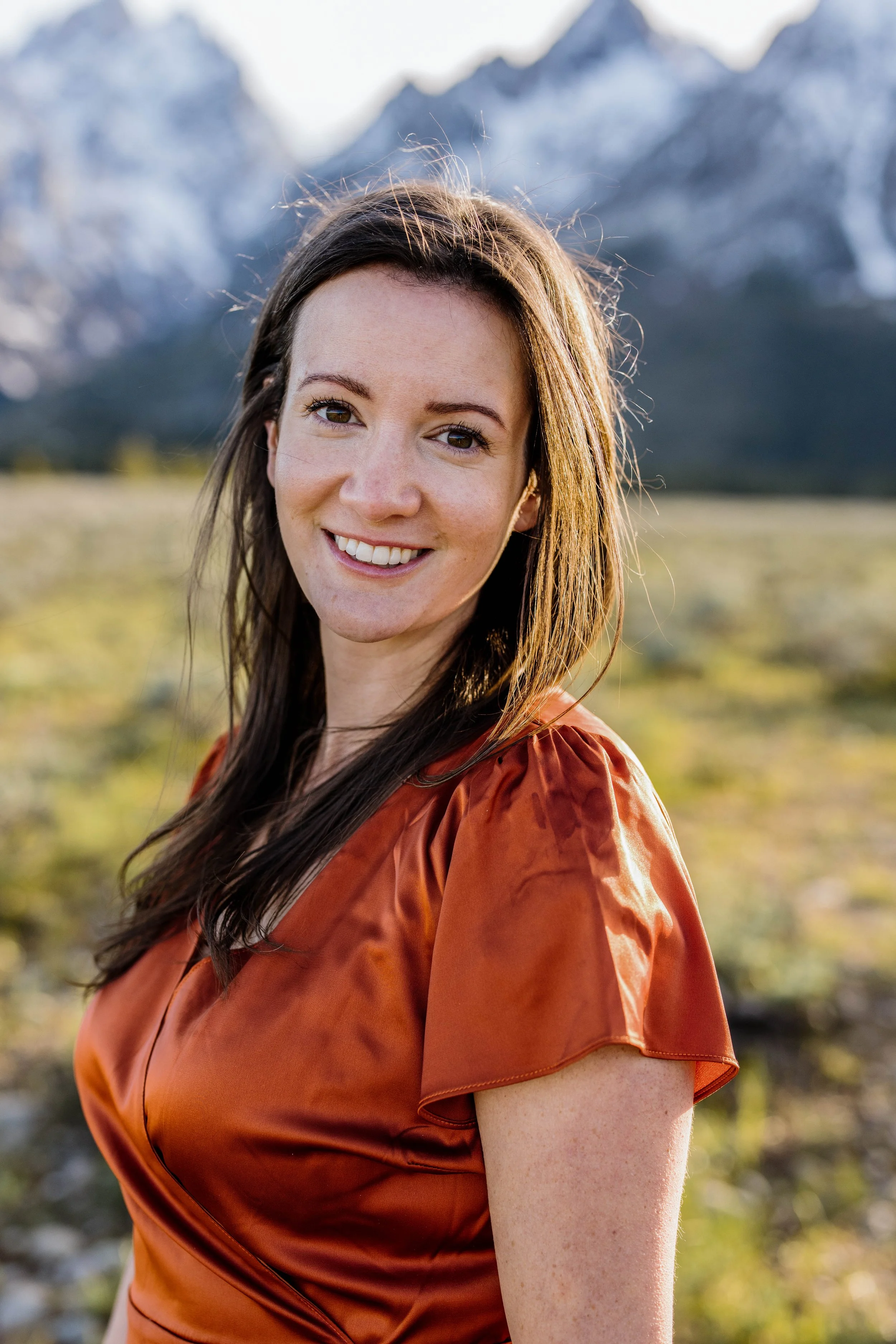A woman with long brown hair smiling outdoors in front of snowy mountains and green field.