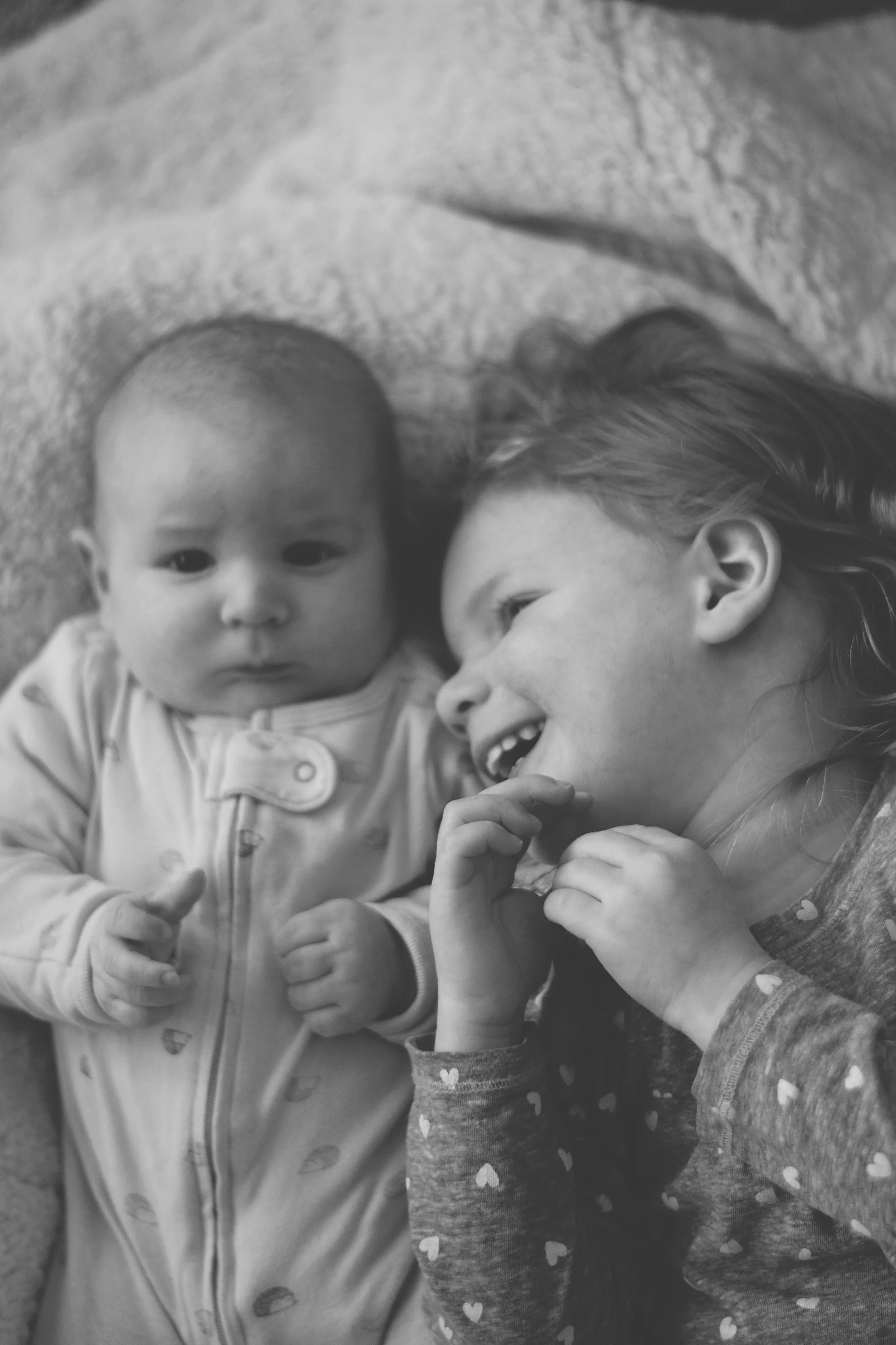 A young girl with long hair shows her teeth and is smiling while looking at a baby with a bald head and big eyes. The baby is facing forward with a neutral expression.