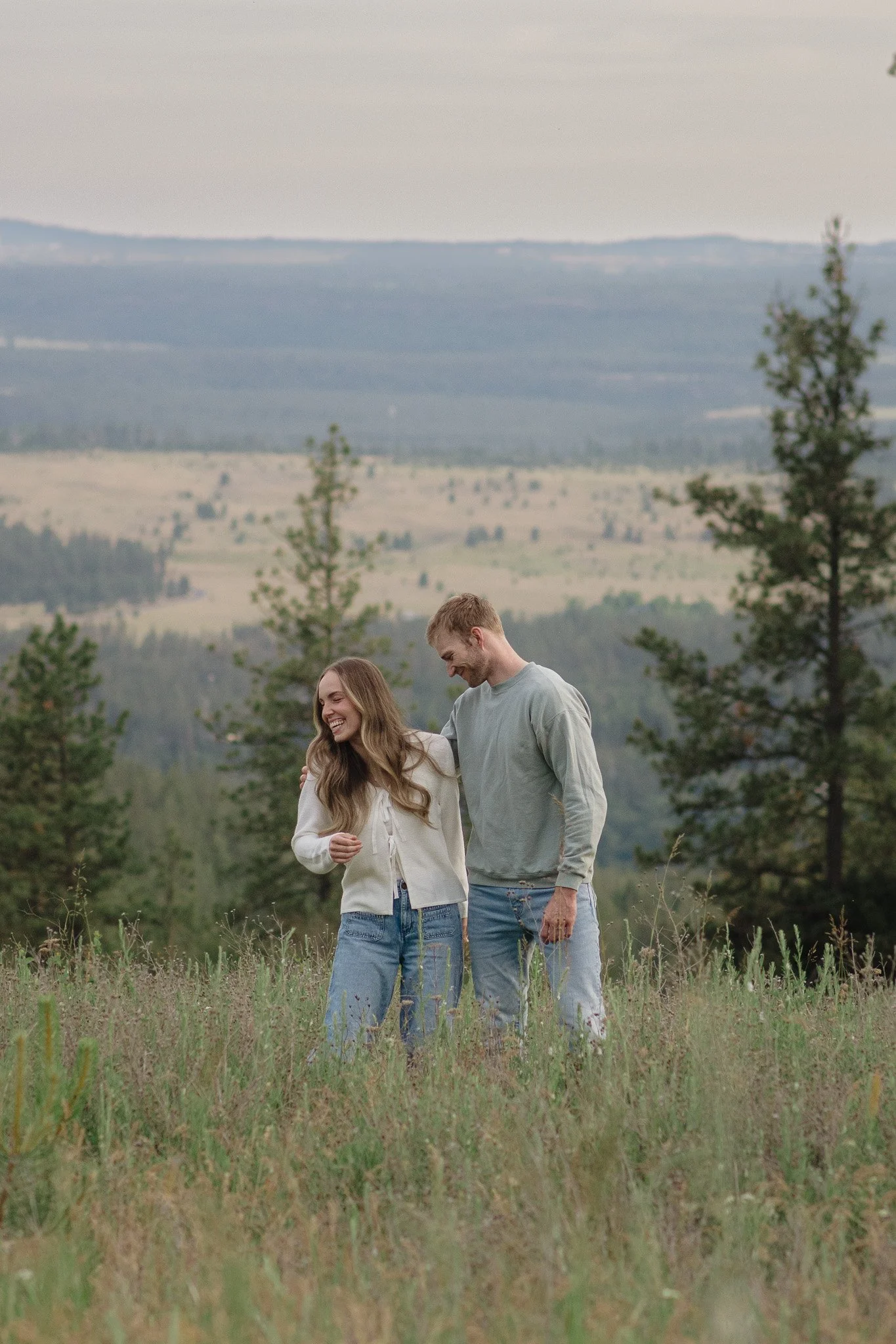 A smiling young woman and a smiling young man walk through a grassy field in Colbert, Washington surrounded by trees, with rolling hills and mountains in the background.
