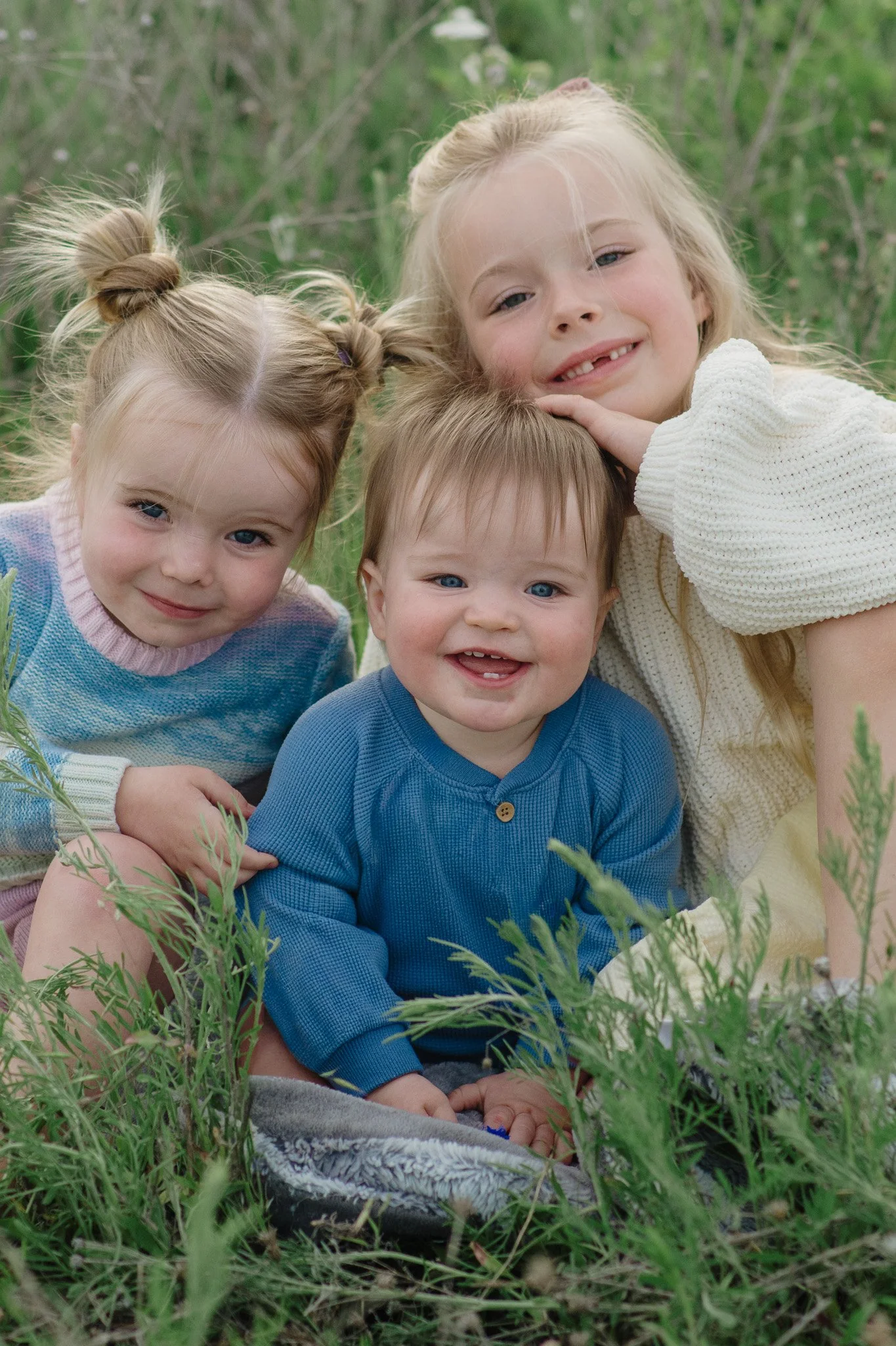 Four children lying close together outdoors among tall grass and greenery, smiling and looking at the camera.