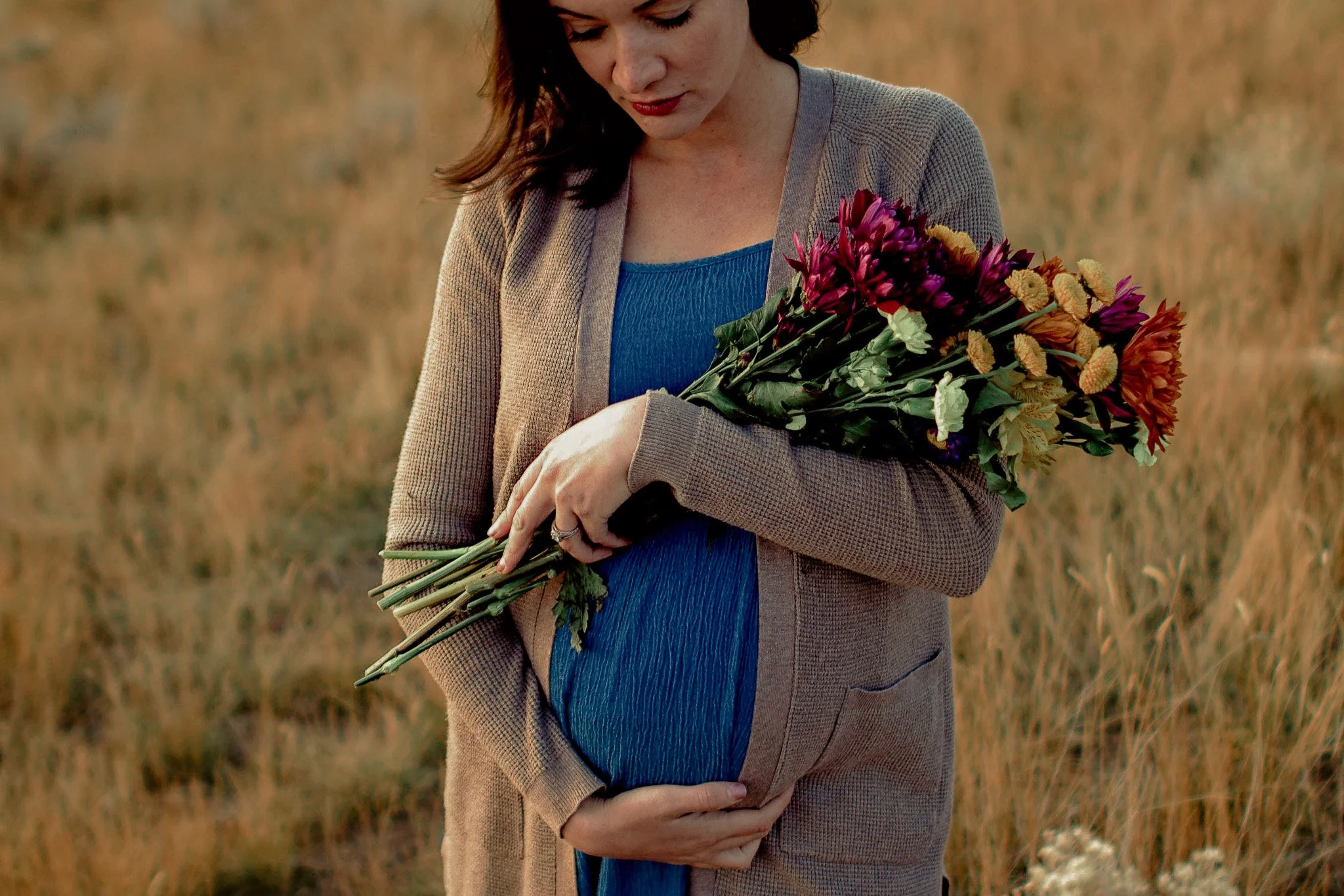A woman standing outdoors in a field holding a bouquet of colorful flowers, with her head slightly bowed and her hand on her pregnant belly.