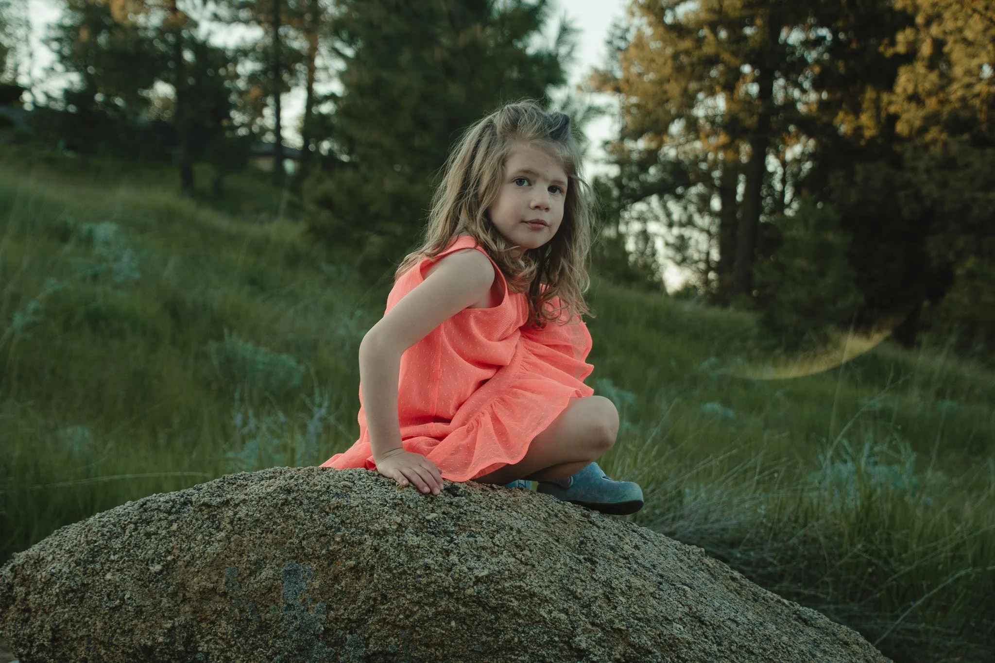 Young girl with long curly hair wearing a coral dress sitting on a rock in a forested outdoor setting