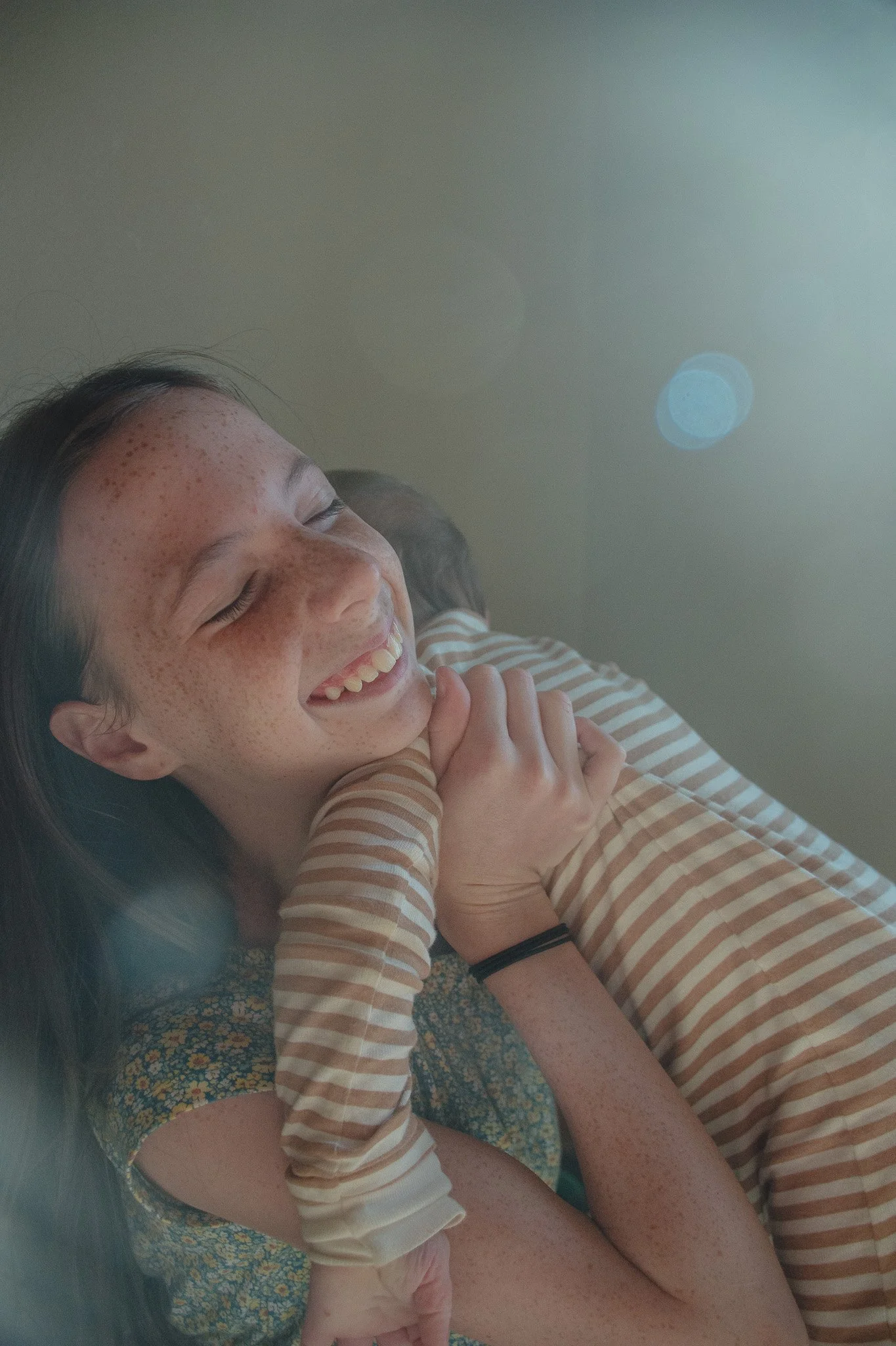 A woman and a small child lying on the floor, smiling with their eyes closed. The woman has long dark hair, freckles, and is wearing a striped shirt. The child, wearing a striped shirt and floral pants, is laying on her chest, with her arms wrapped a