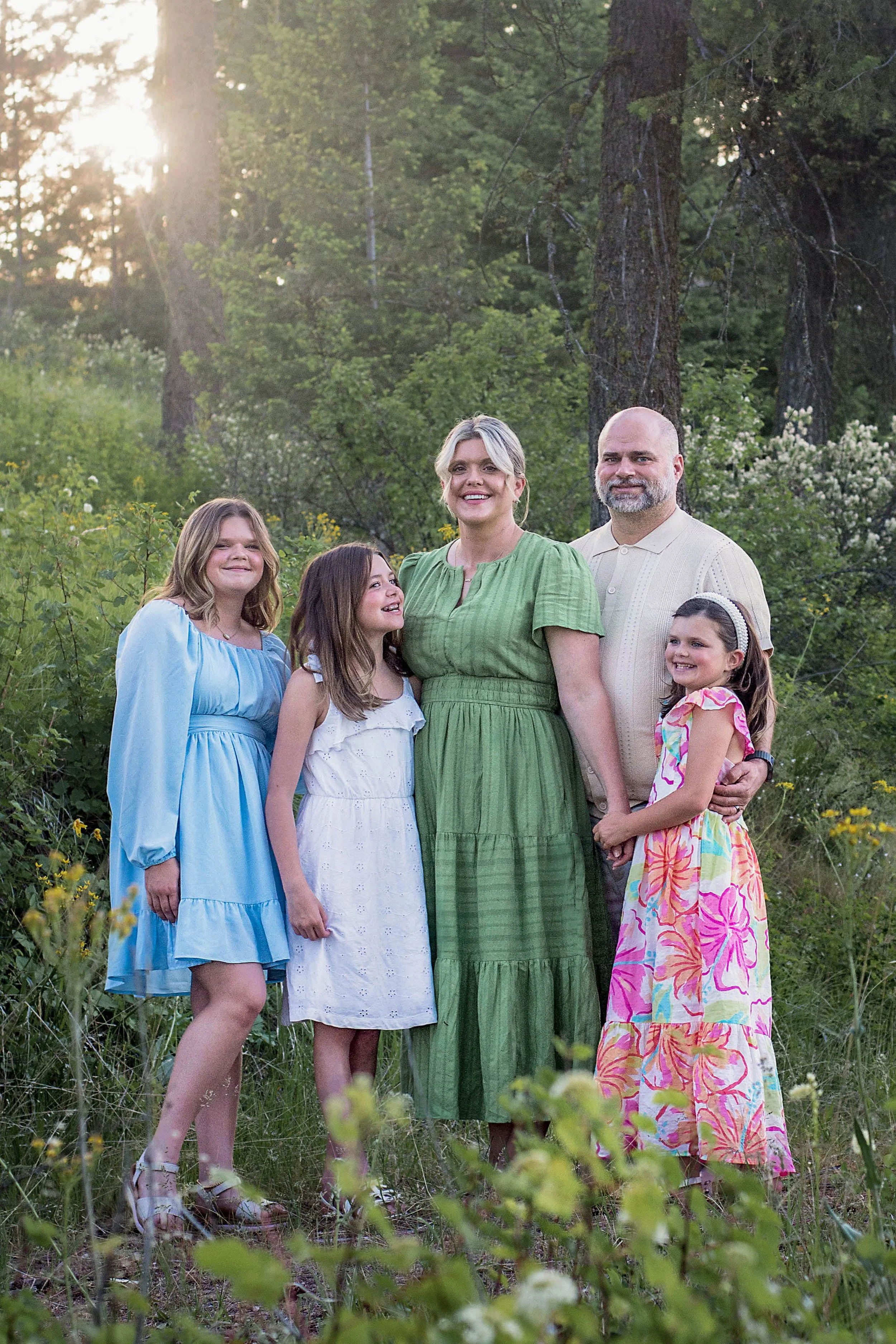 A family of six posing outdoors in a lush green forest. The group includes three adult women and one man, along with three young girls. They are dressed in colorful spring or summer dresses and are smiling at the camera, with the warm sunlight filter