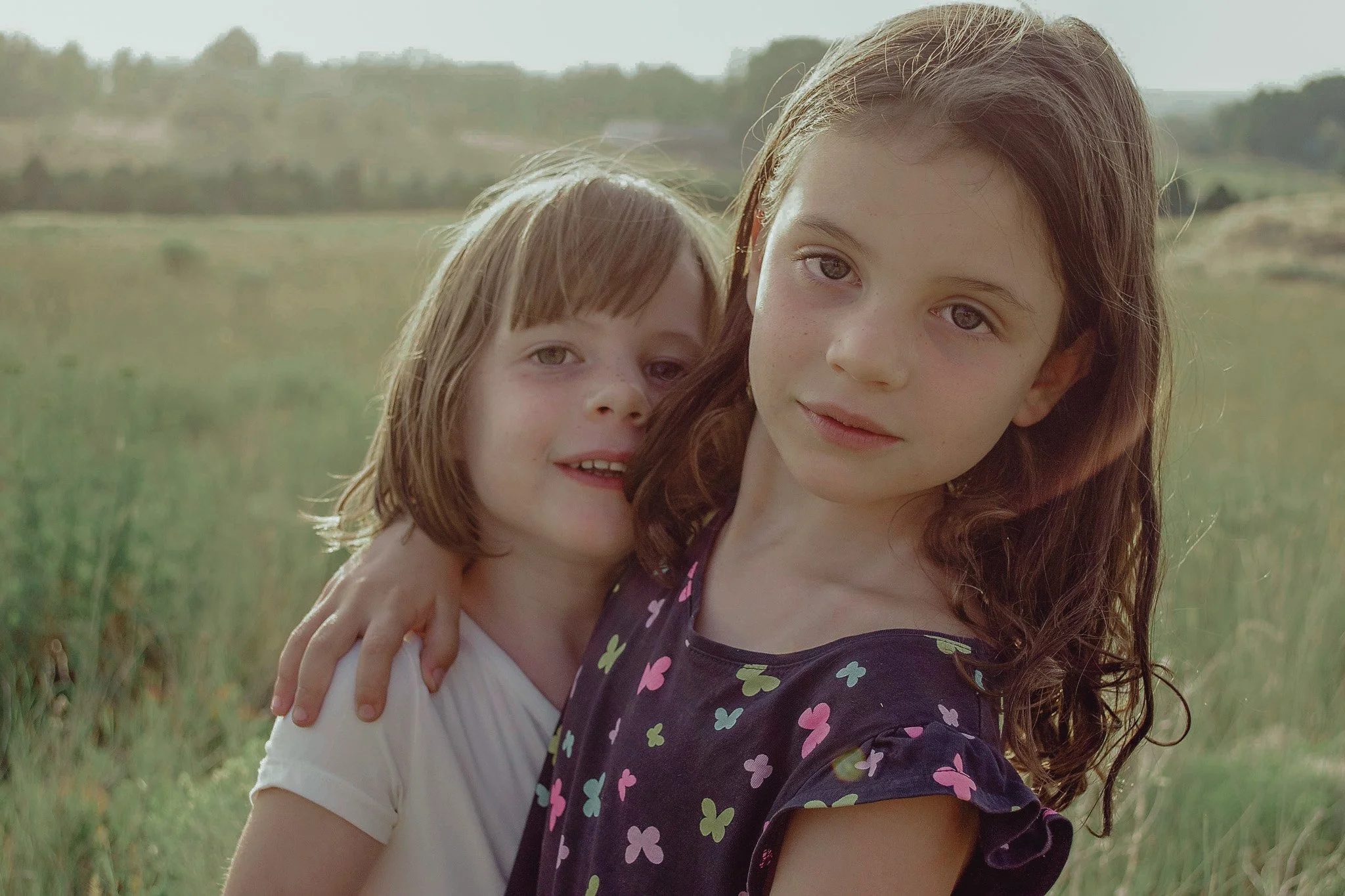 Two young girls with light skin and brown hair standing in a grassy field, embracing each other and smiling softly on a sunny day with a blurred tree line in the background.