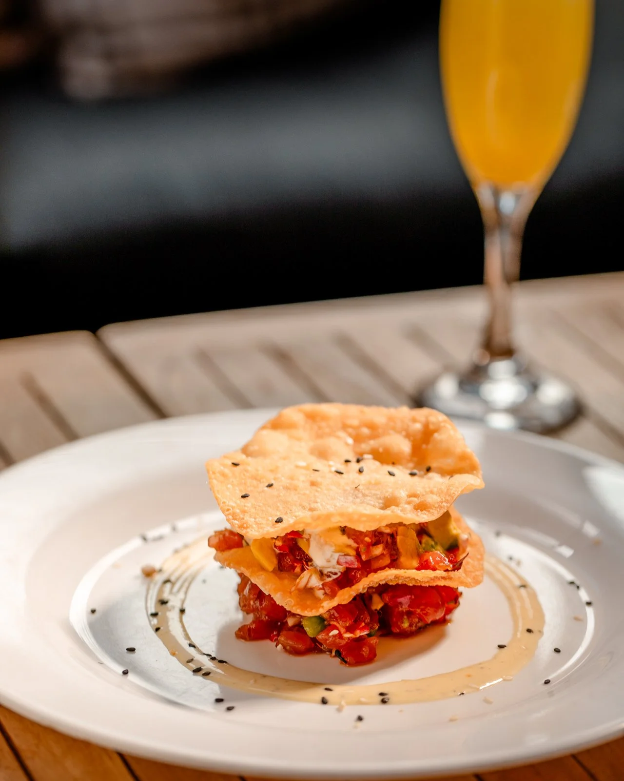 A gourmet dish with layered ingredients topped with a crispy wafer on a white plate, with black sesame seeds and sauce. A drink with an orange-colored beverage in a tall glass is in the background.