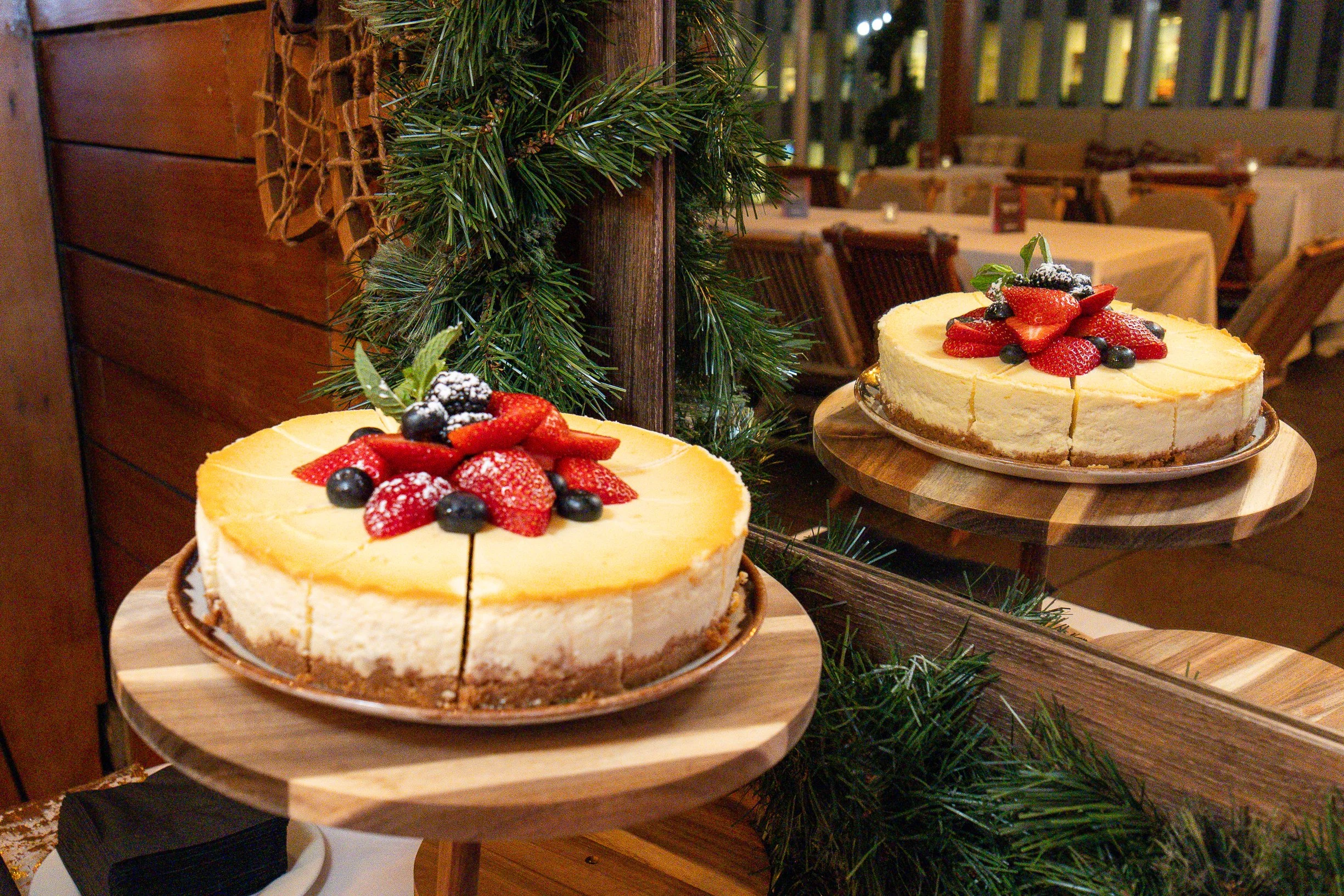 Cheesecake topped with strawberries, blueberries, and mint leaves on a wooden stand at a restaurant decorated with green garland.