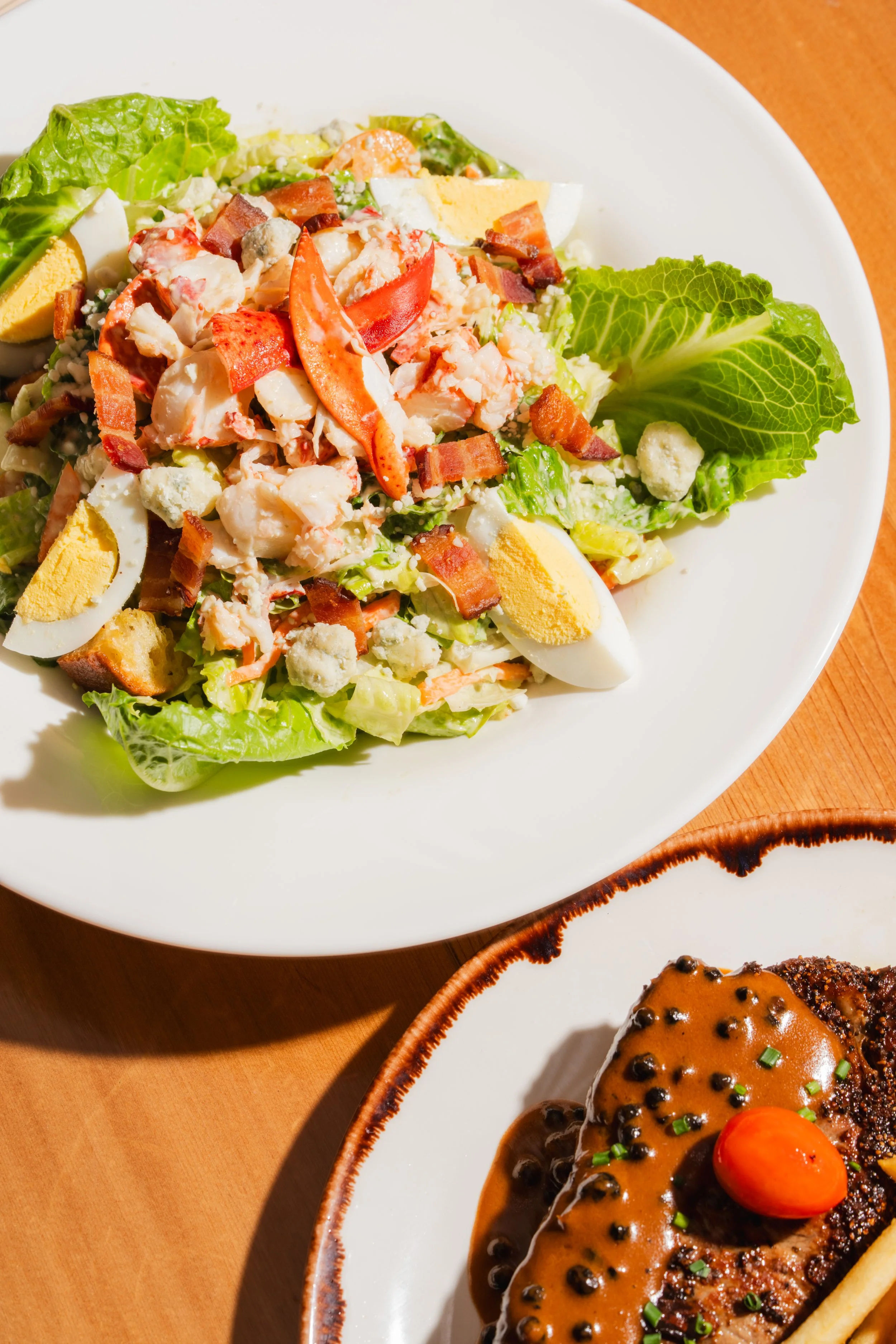 Close-up of a plate with a salad, boiled eggs, bacon bits, and shredded chicken. Part of another dish with cooked meat and sauce is visible on the side.