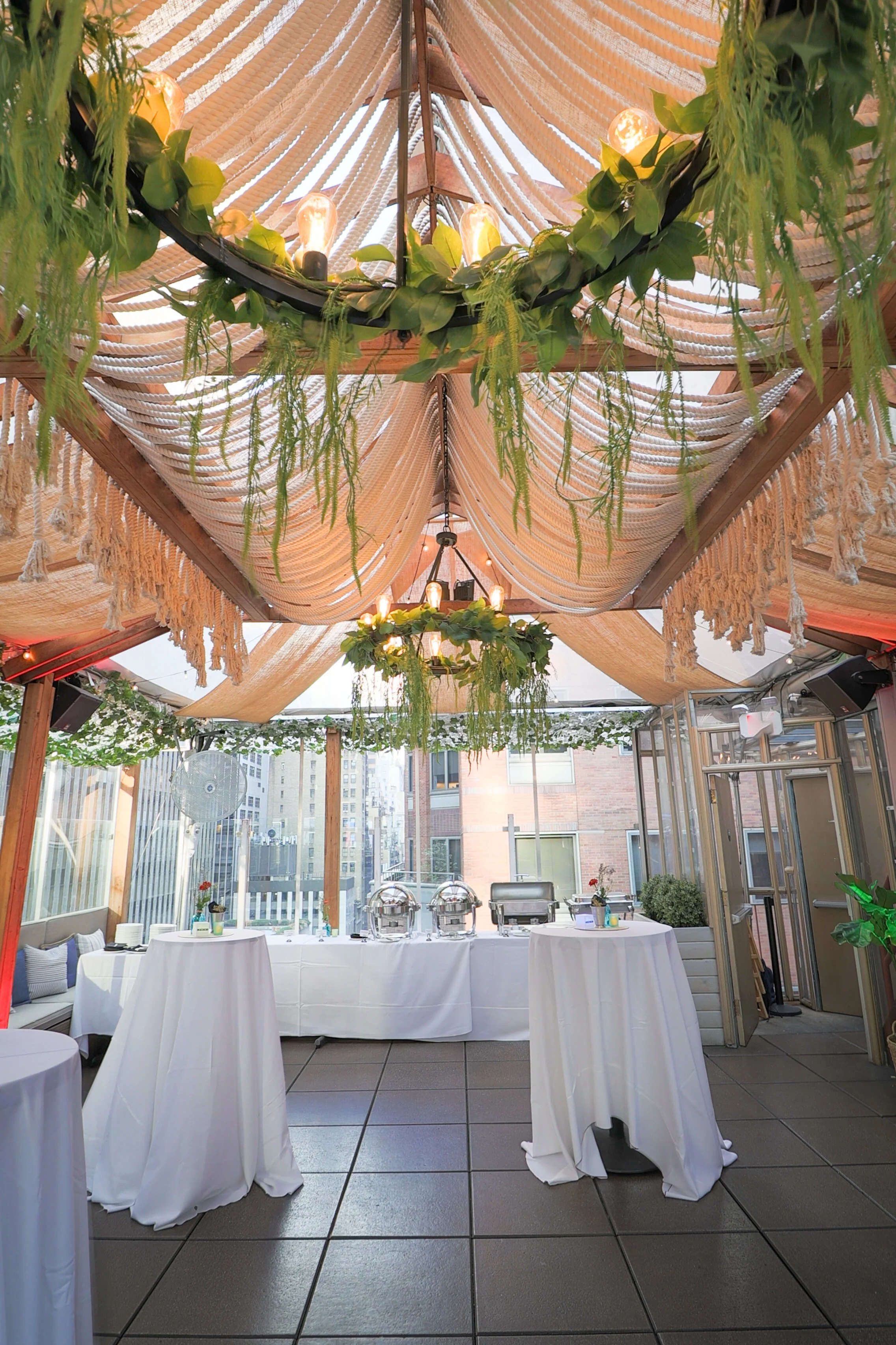 Outdoor event space with a ceiling decorated with beige fabric drapes, greenery, and hanging lights. There are round tables with white tablecloths and food serving stations in the background. The space is enclosed with glass walls showing urban build