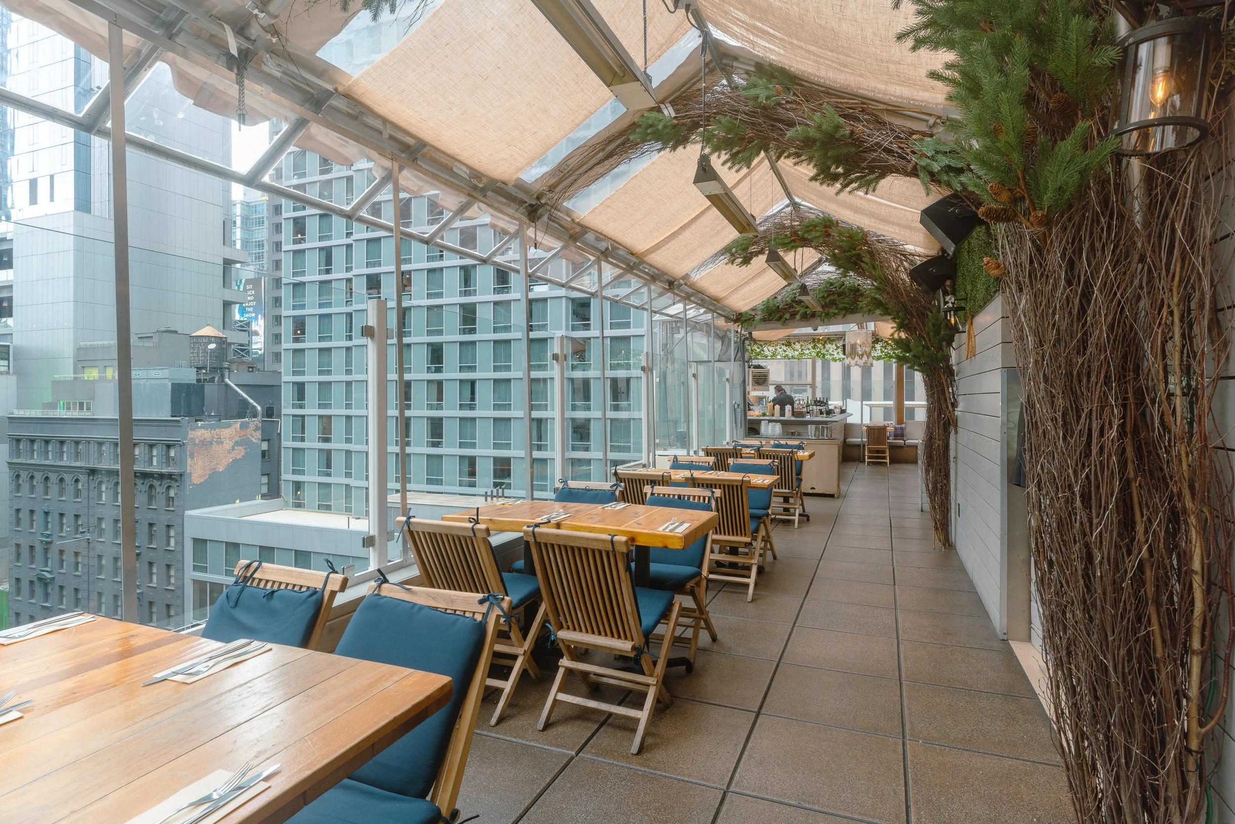 A rooftop restaurant or bar with wooden tables and blue cushioned chairs, decorated with green pine branches and twigs along the wall, and overlooking city skyscrapers through large glass windows.