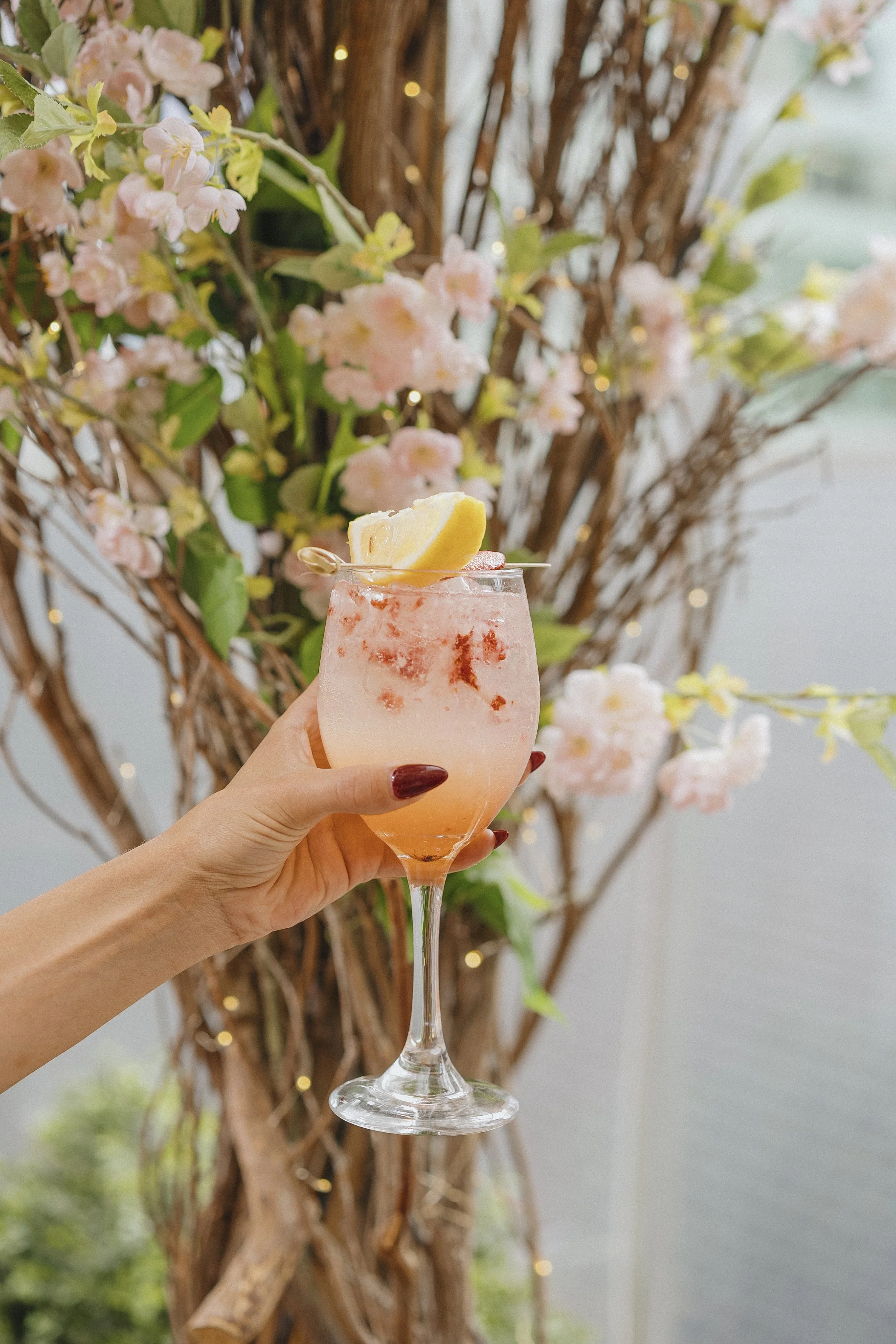 A hand holding a glass of pink cocktail with a lemon wedge garnish, in front of a flower arrangement with pink flowers and twigs.