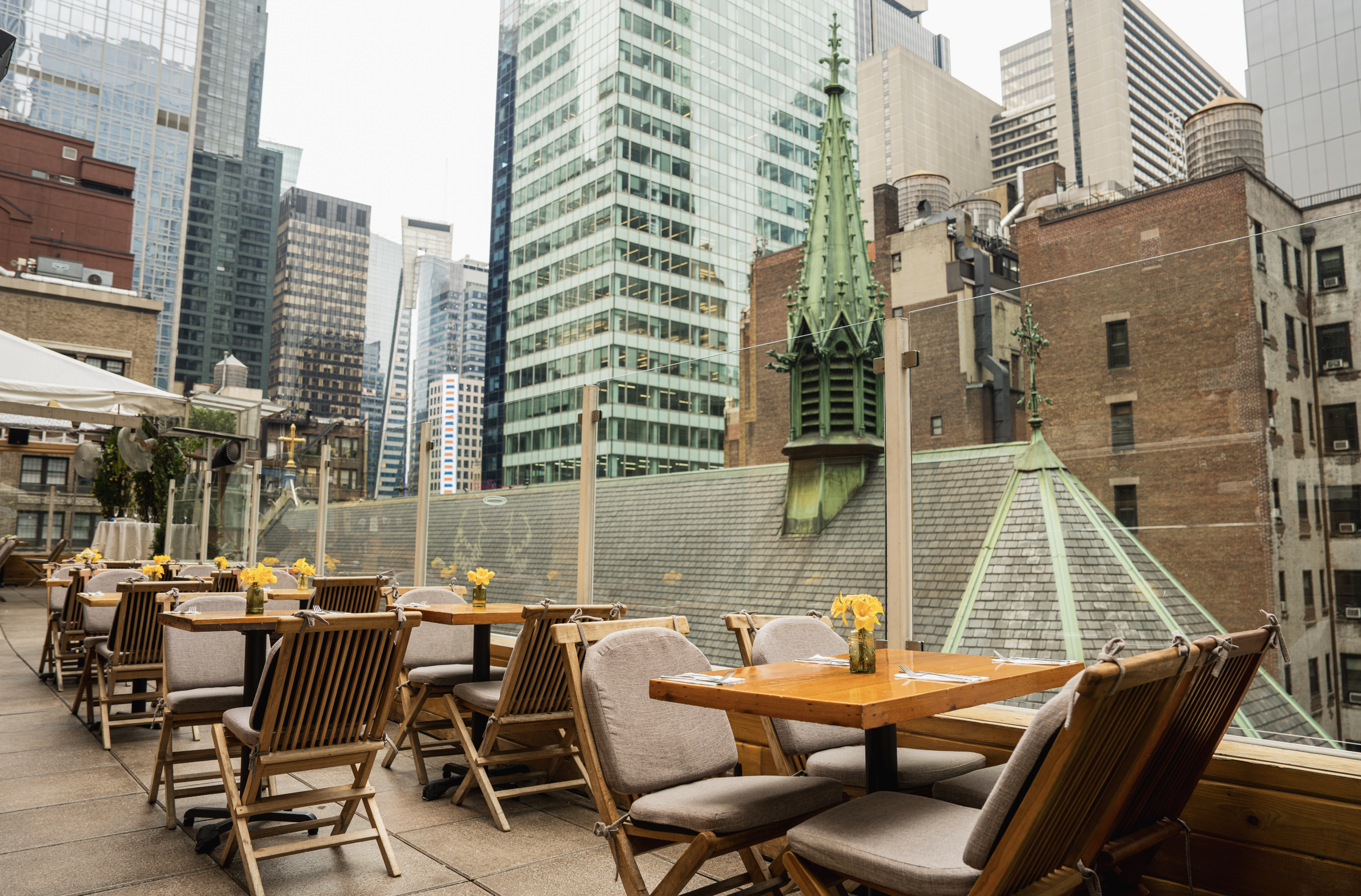 Outdoor rooftop dining area with wooden tables and beige cushioned chairs, decorated with yellow flowers in glass vases. In the background, tall modern glass skyscrapers and older brick buildings create an urban cityscape.