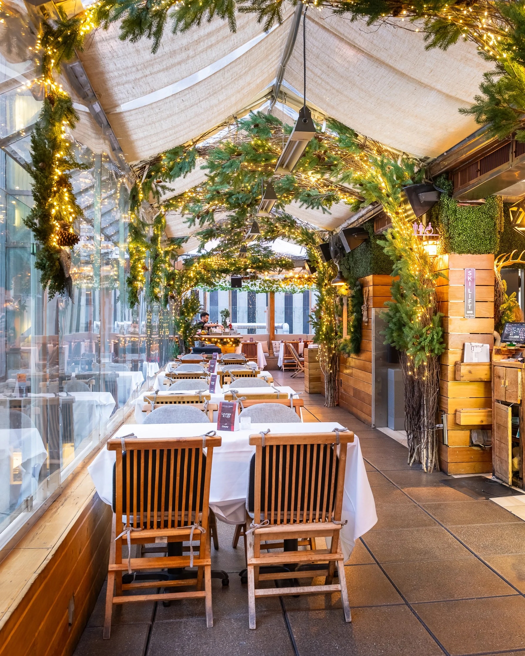 A restaurant with wooden tables and chairs, decorated for Christmas with green garlands and string lights hanging from the ceiling. There is a glass wall on the left and a waiter serving customers in the background.