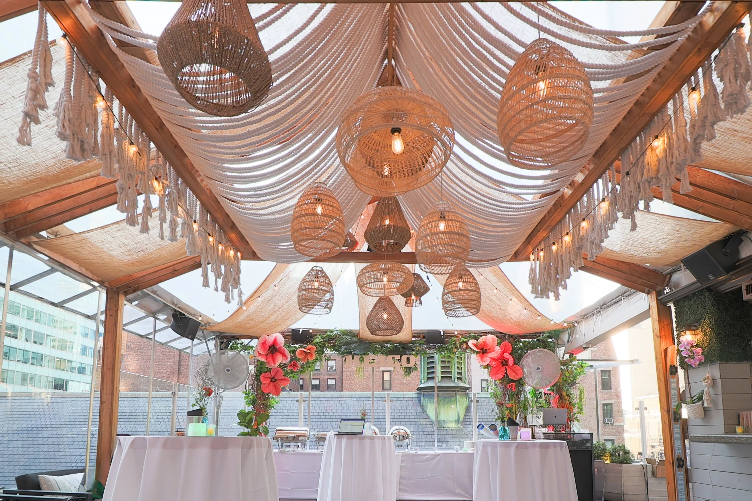 Decorative indoor space with hanging woven lantern-style lights, draped white fabric with tassels, string lights, and potted pink flowers, set in a space with large windows showing city buildings.