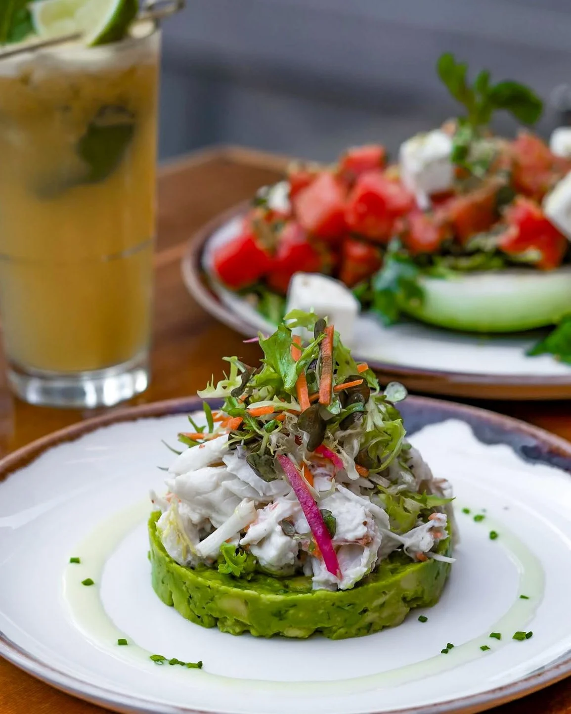 A gourmet seafood salad on a white plate, served on a bed of mashed avocado, topped with mixed microgreens and shredded vegetables, with a drizzle of dressing. In the background, there are plates of chopped tomato and cucumber salad and a glass of ic