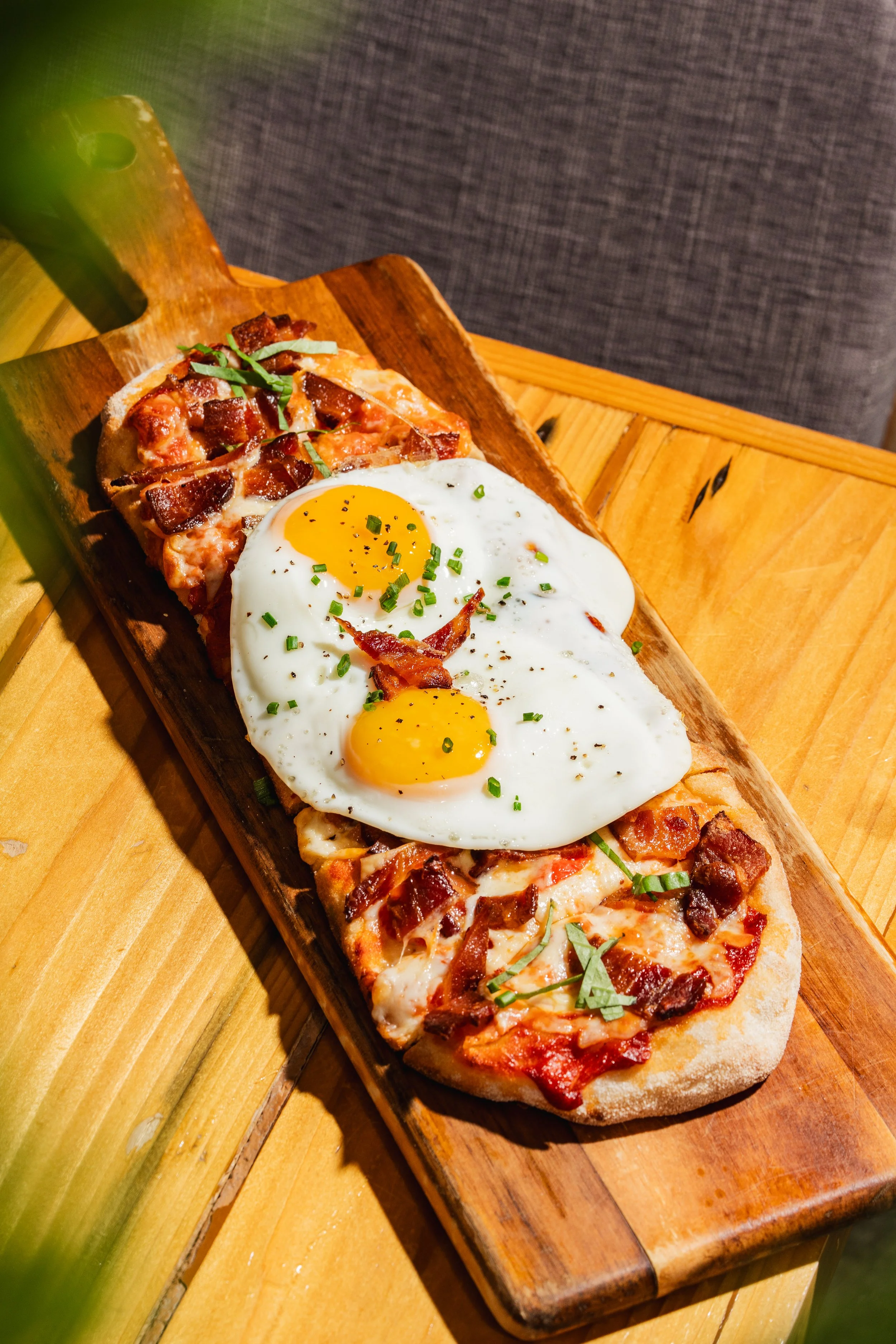 Pizza topped with bacon, eggs, and green herbs on a wooden serving board.