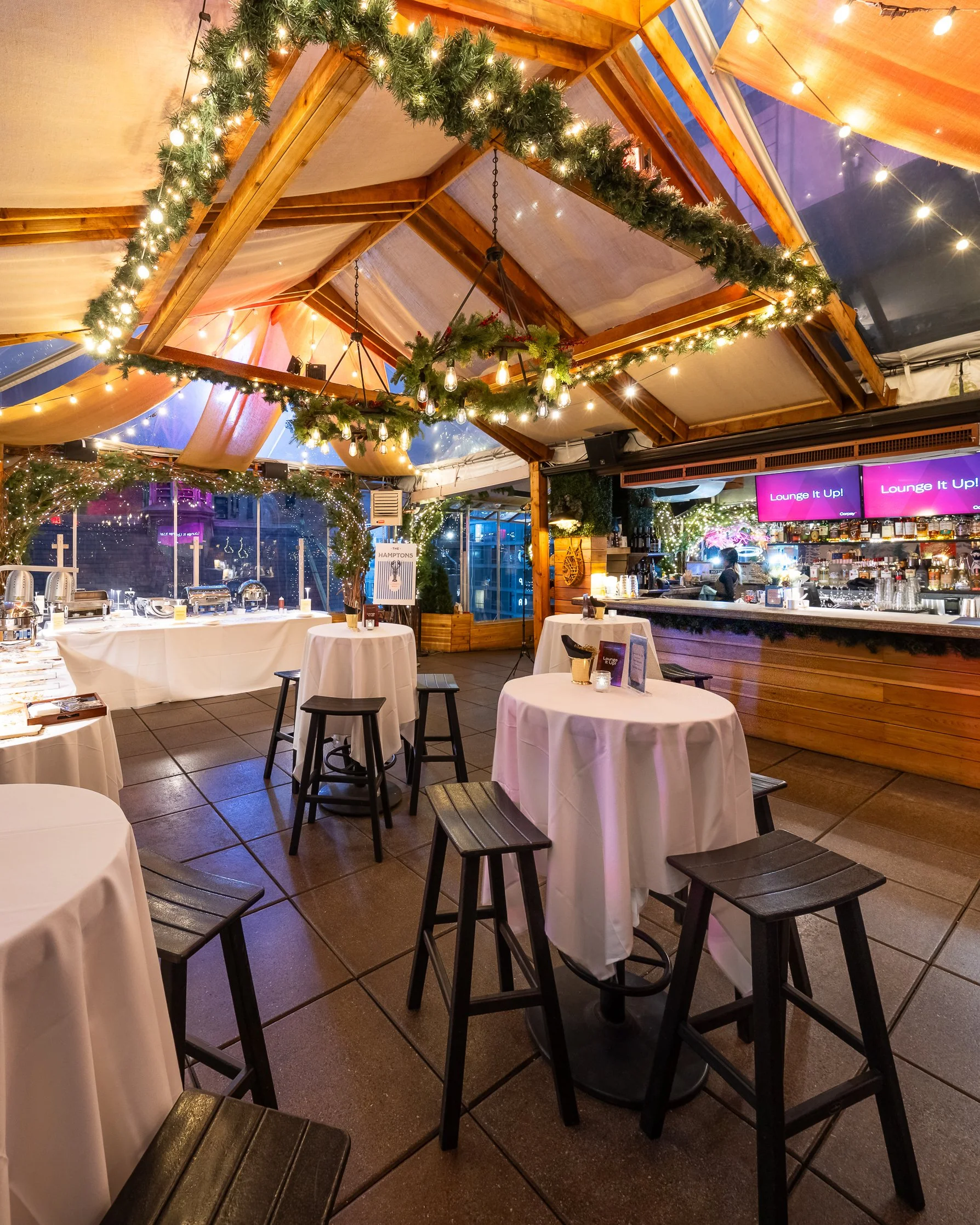 Indoor restaurant or bar with tables covered in white tablecloths, topped with small candles, black chairs, and a bar area in the background. Decor includes string lights, greenery, and wood accents, creating a festive atmosphere.