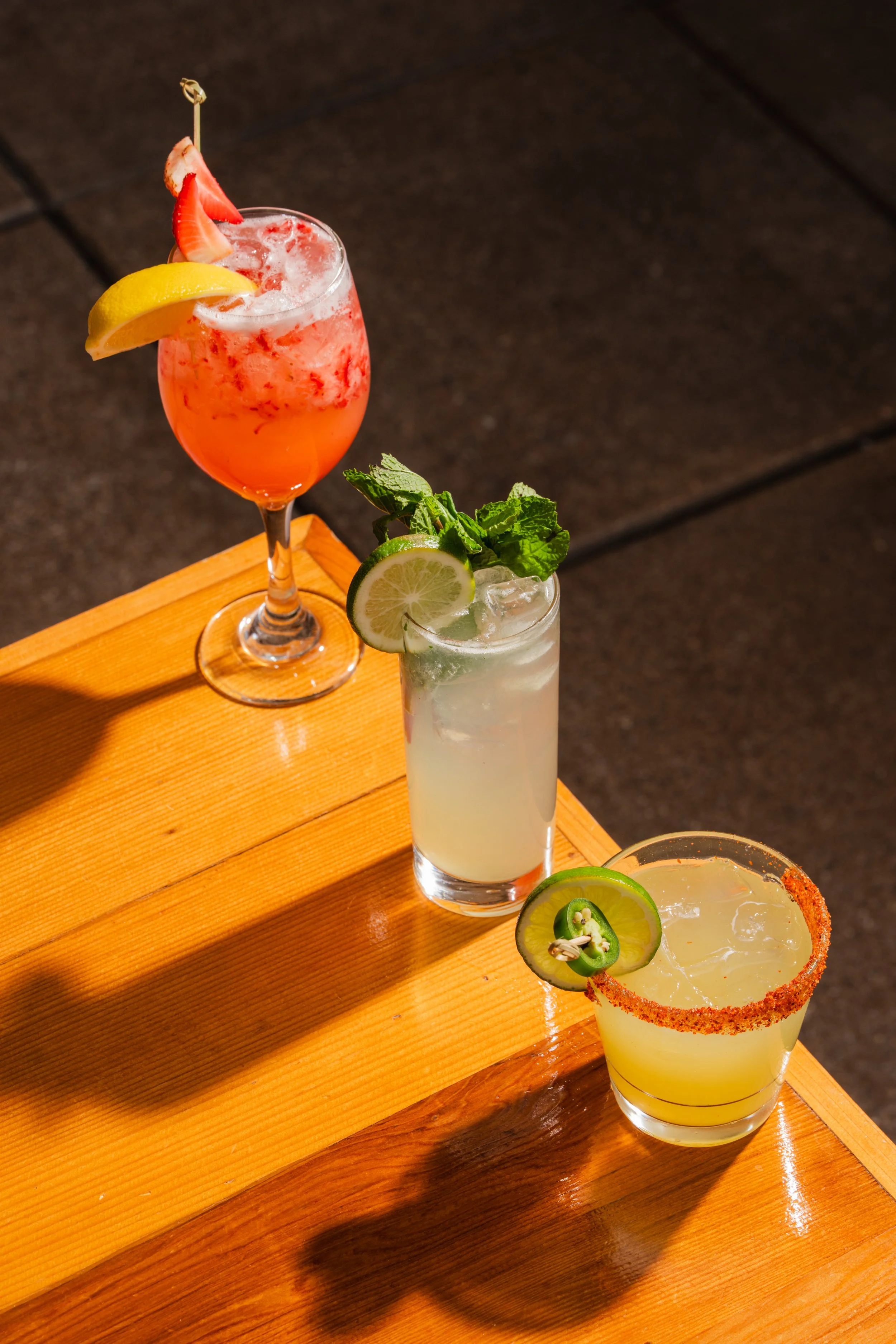 Three colorful cocktails garnished with fruit slices and herbs on a wooden table.