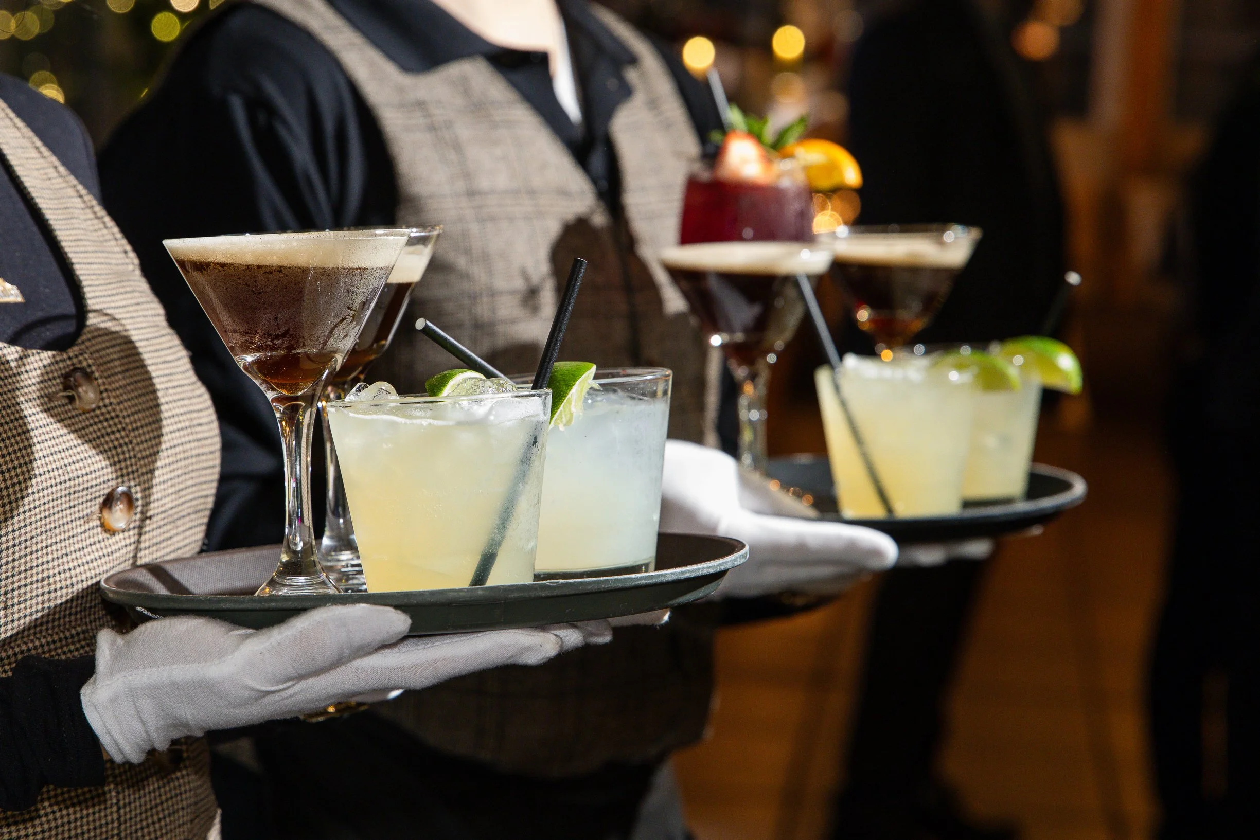 A server in white gloves carries a tray with several cocktails garnished with lime and fruit slices, set against a dimly lit background with blurred warm lights.