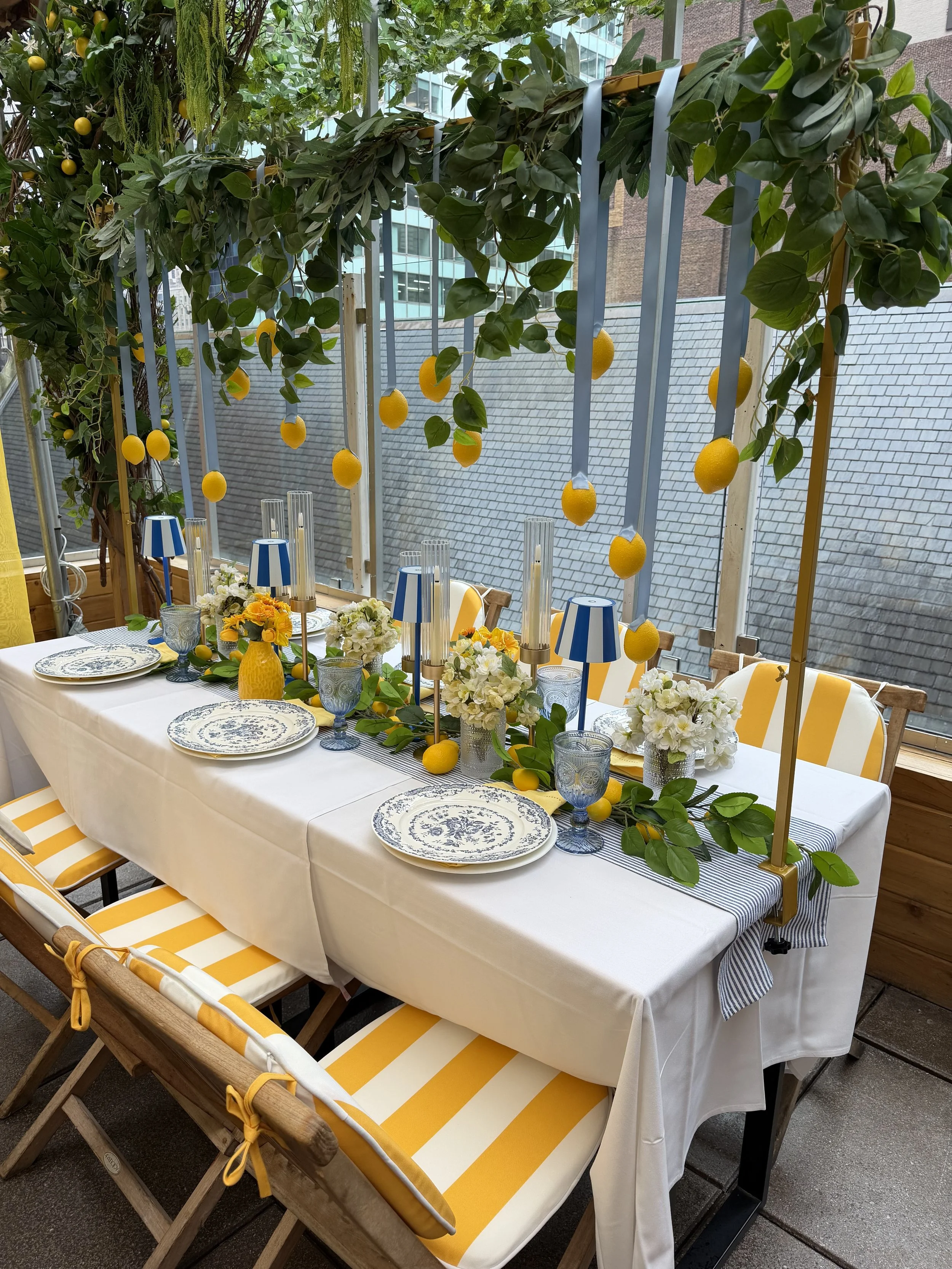 A decorated dining table with a white tablecloth, colorful tableware including plate and glasses, surrounded by yellow and white striped cushions, and adorned with floral arrangements and lemon-themed decorations suspended from a leafy green overhead structure with ribbons.