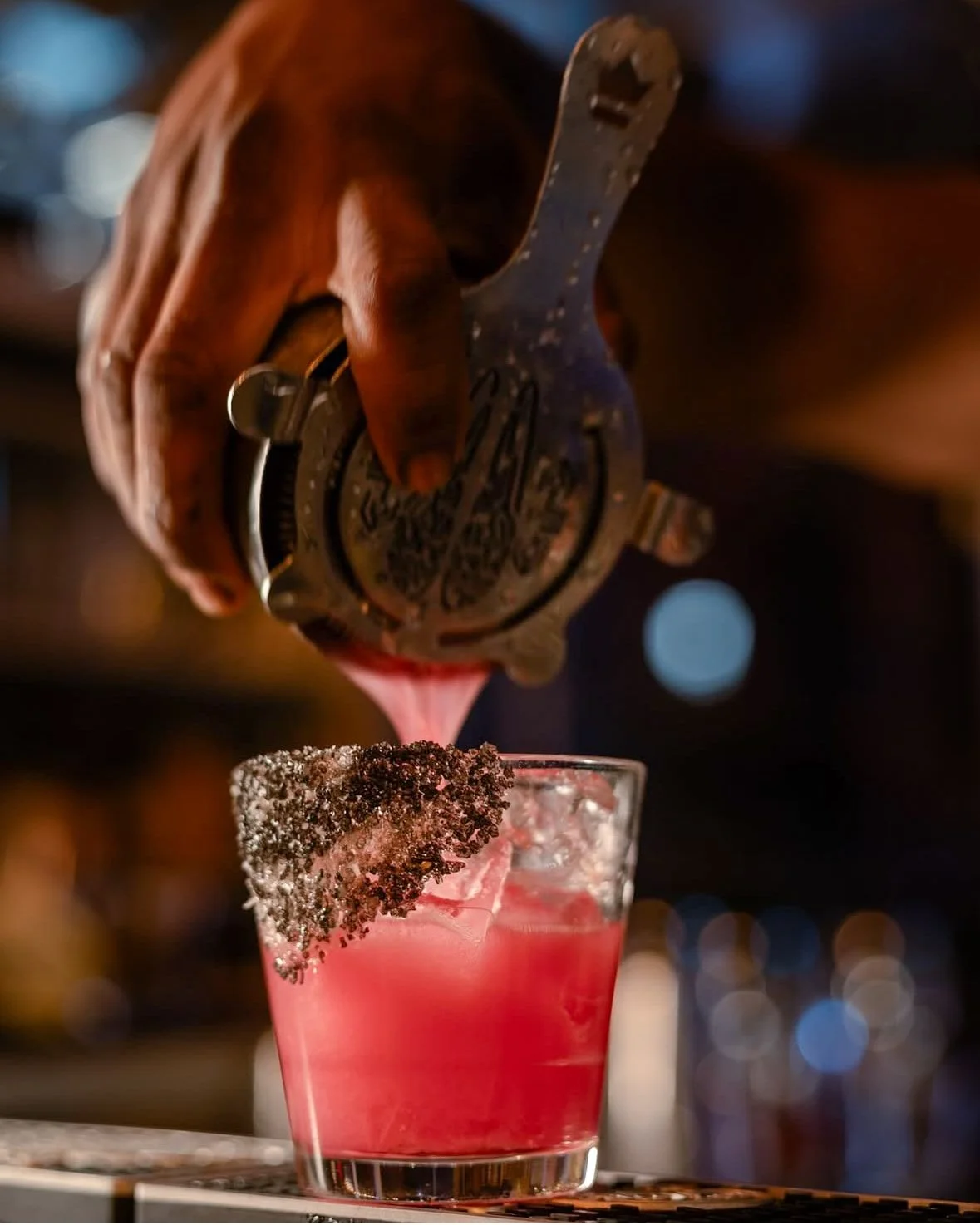 A person is pouring a pink cocktail from a metal shaker into a glass with a black rim and pink sugar rim, on a bar counter with bokeh lights in the background.
