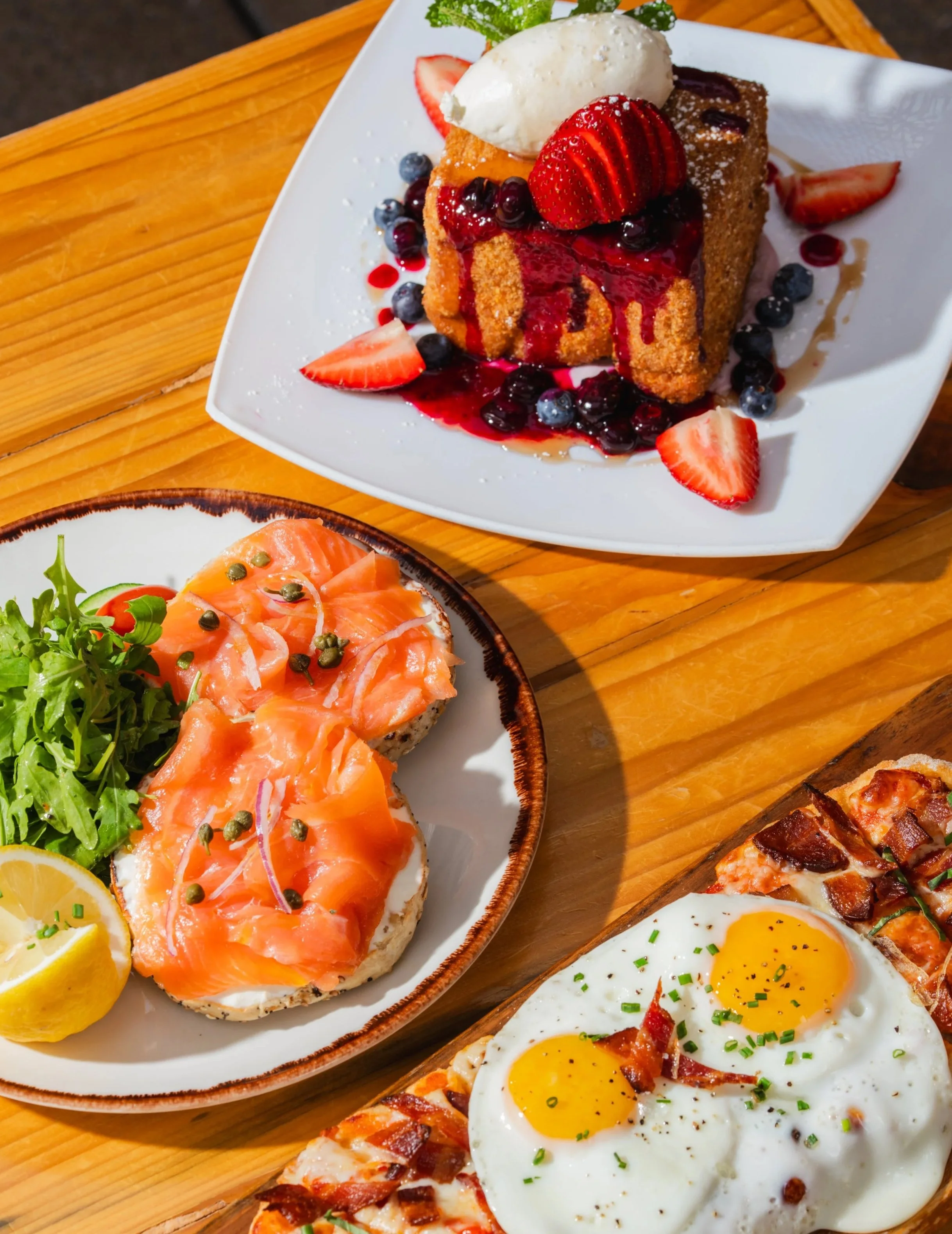 A dessert plate with a slice of cake topped with strawberries, blueberries, and a scoop of vanilla ice cream, drizzled with berry syrup, served with lemon slices and greens; a breakfast plate with smoked salmon on bread garnished with capers and lemon wedges; a breakfast plate with fried eggs, bacon, and hash browns.