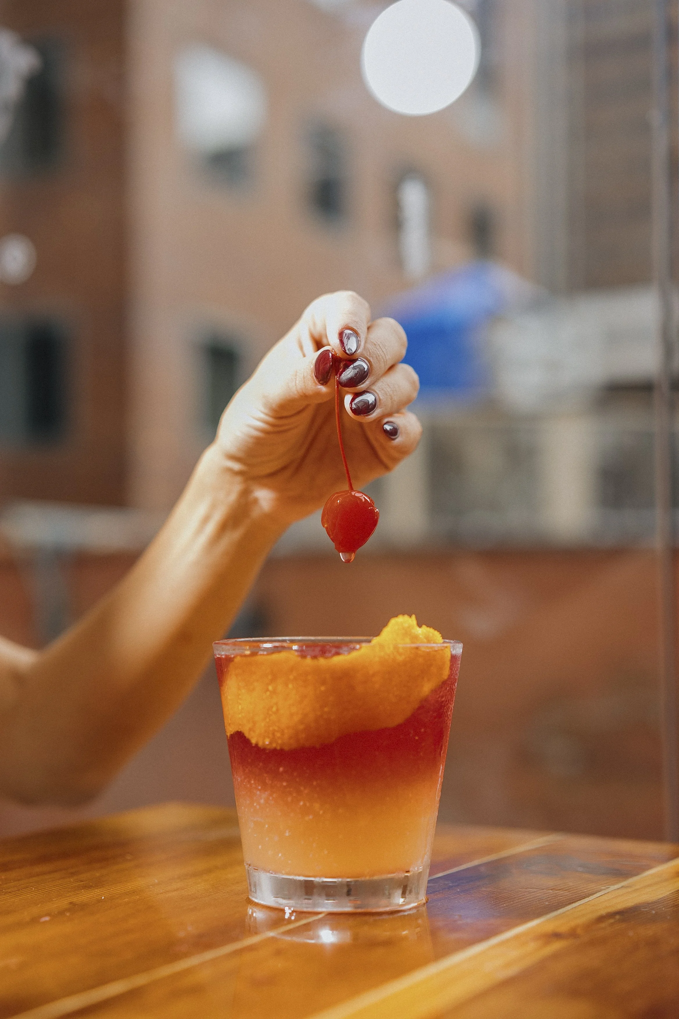 A hand with painted nails holds a maraschino cherry above a brightly colored cocktail with a slice of fruit on the rim, on a wooden table, with a blurred background of buildings.