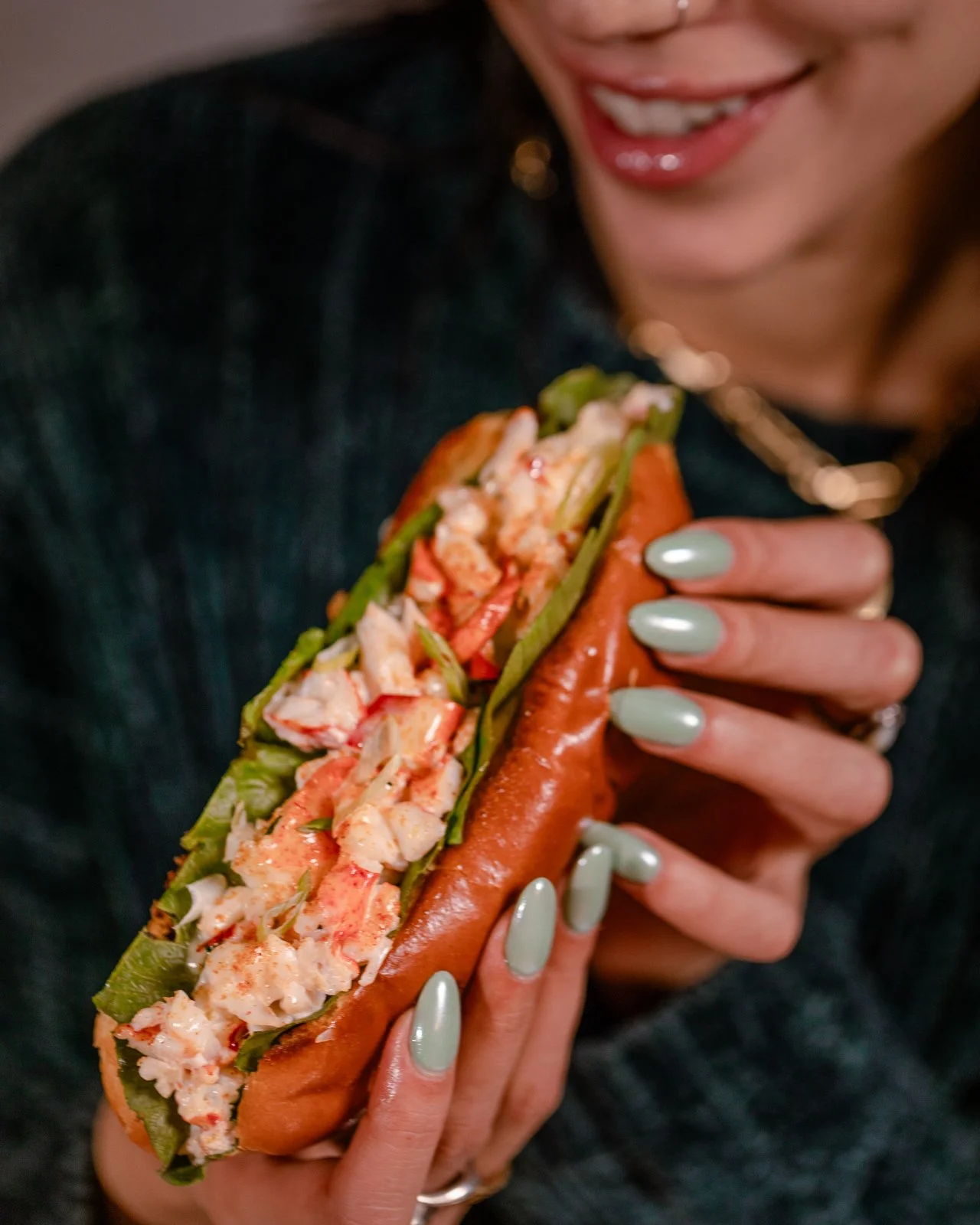 A woman with mint green nail polish holding a lobster roll with lettuce, lobster meat, and a hot dog bun.